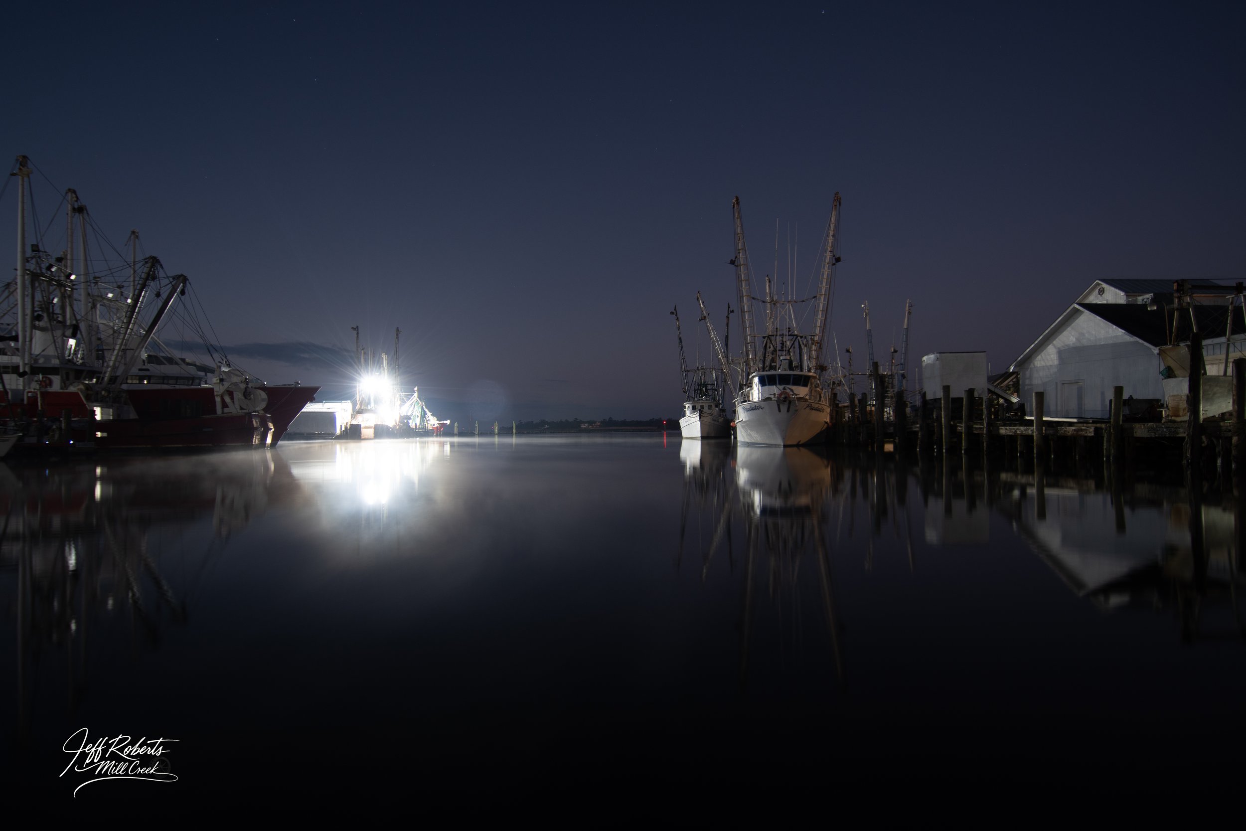 Night scene of boats docked at a marina, with their reflections on calm water and a dark sky in the background.