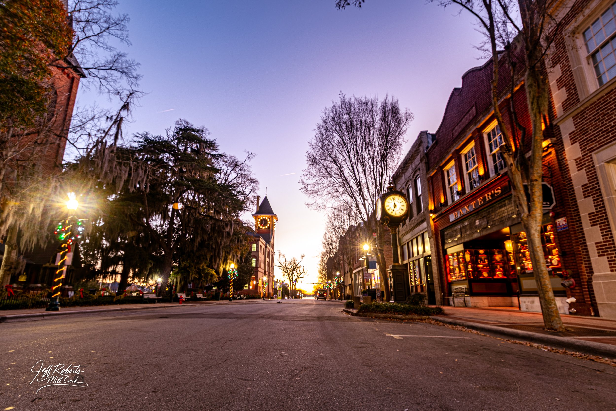 A quiet city street at sunset with buildings decorated for Christmas, including a store called Batters, vintage street clocks, and holiday lights on trees, with a colorful sky in the background.