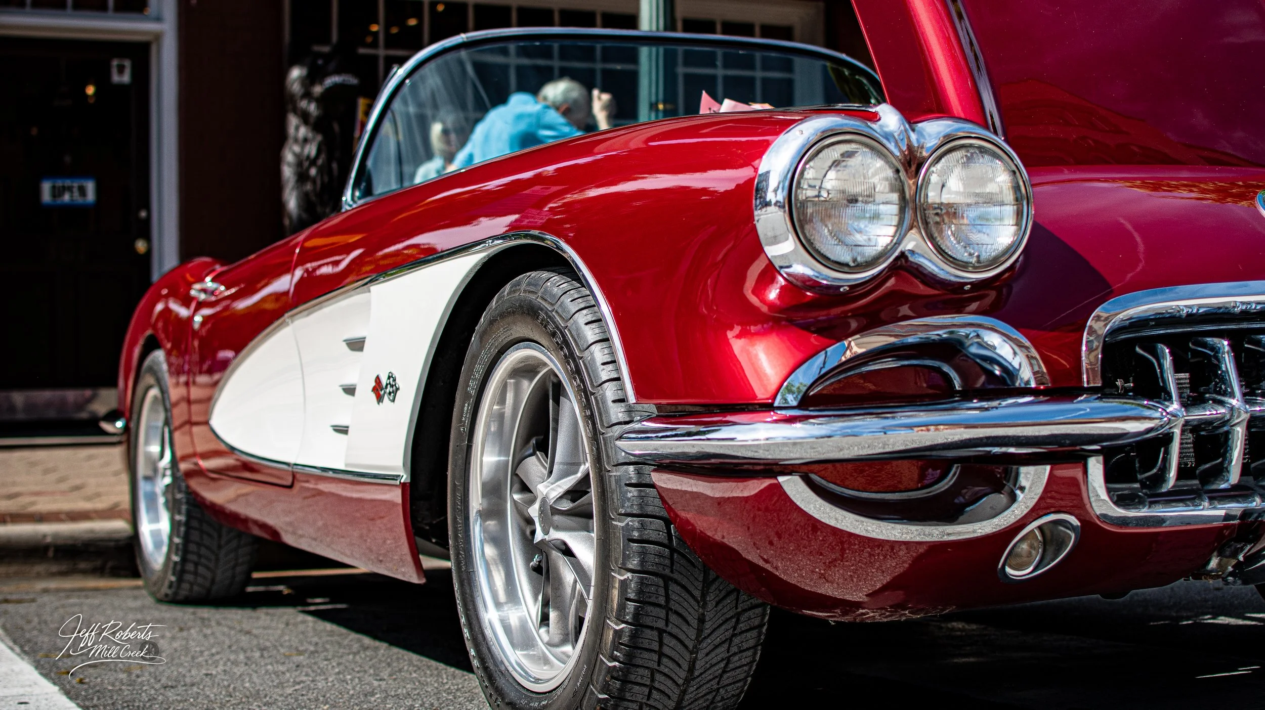 Close-up of a red vintage Chevrolet Corvette with white side panel, chrome detailing, and dual round headlights, parked on street during a car show.