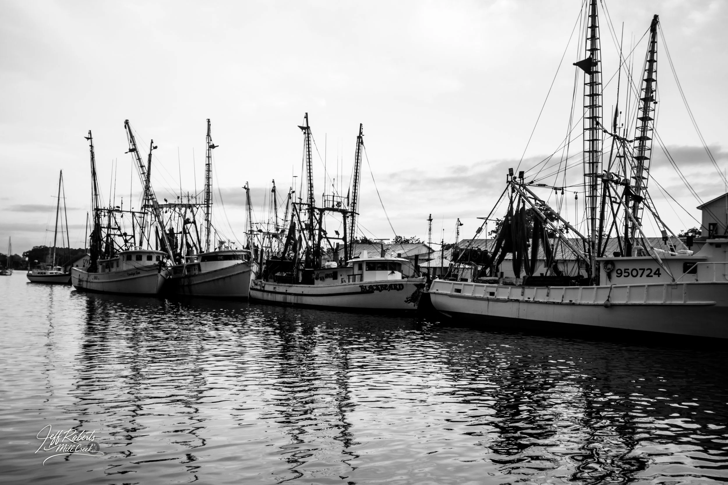 Black and white photo of several fishing boats docked at a marina, with their reflections in the water.