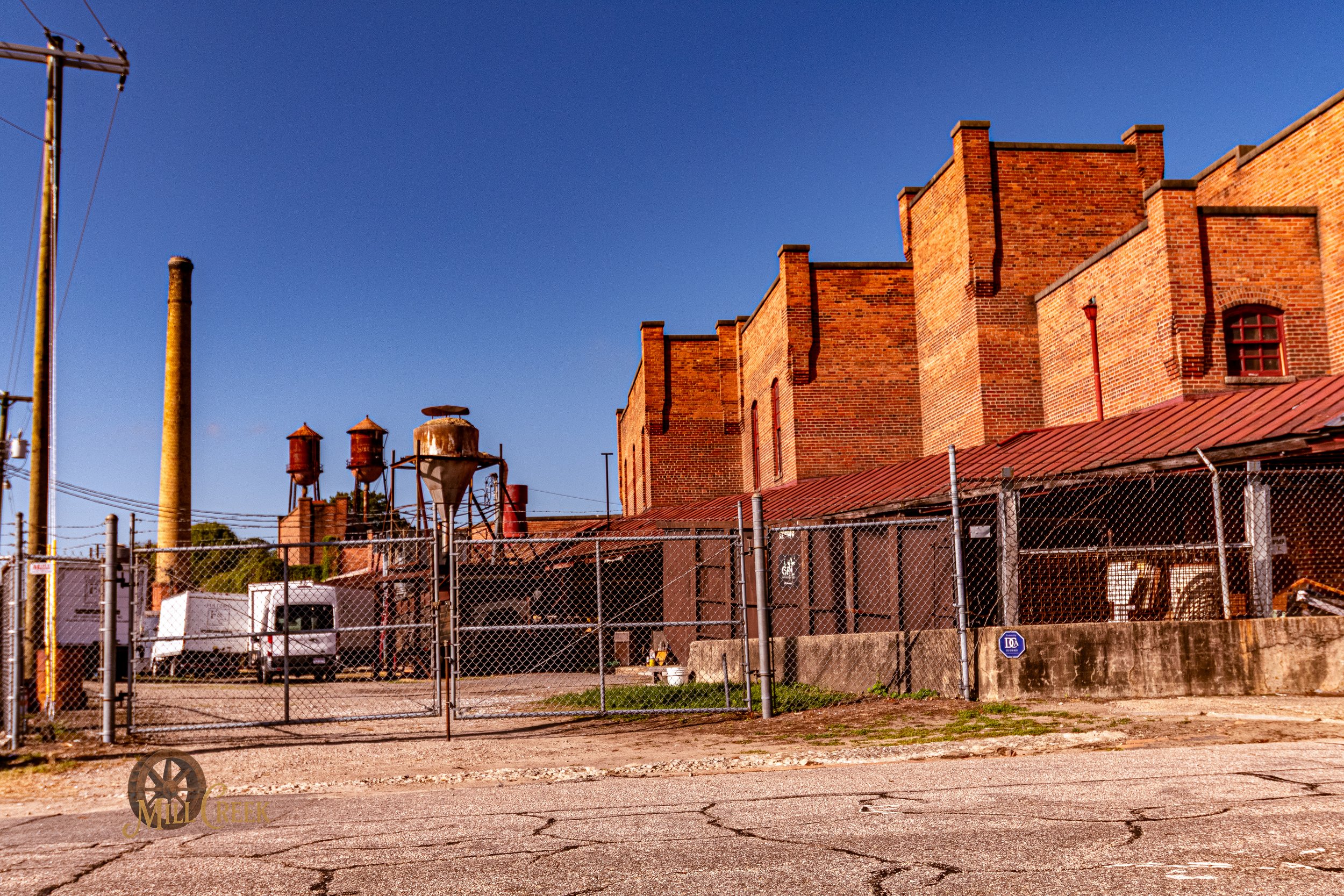 A red brick industrial building with a fenced yard, old water tanks, and a truck parked outside, under a clear blue sky.