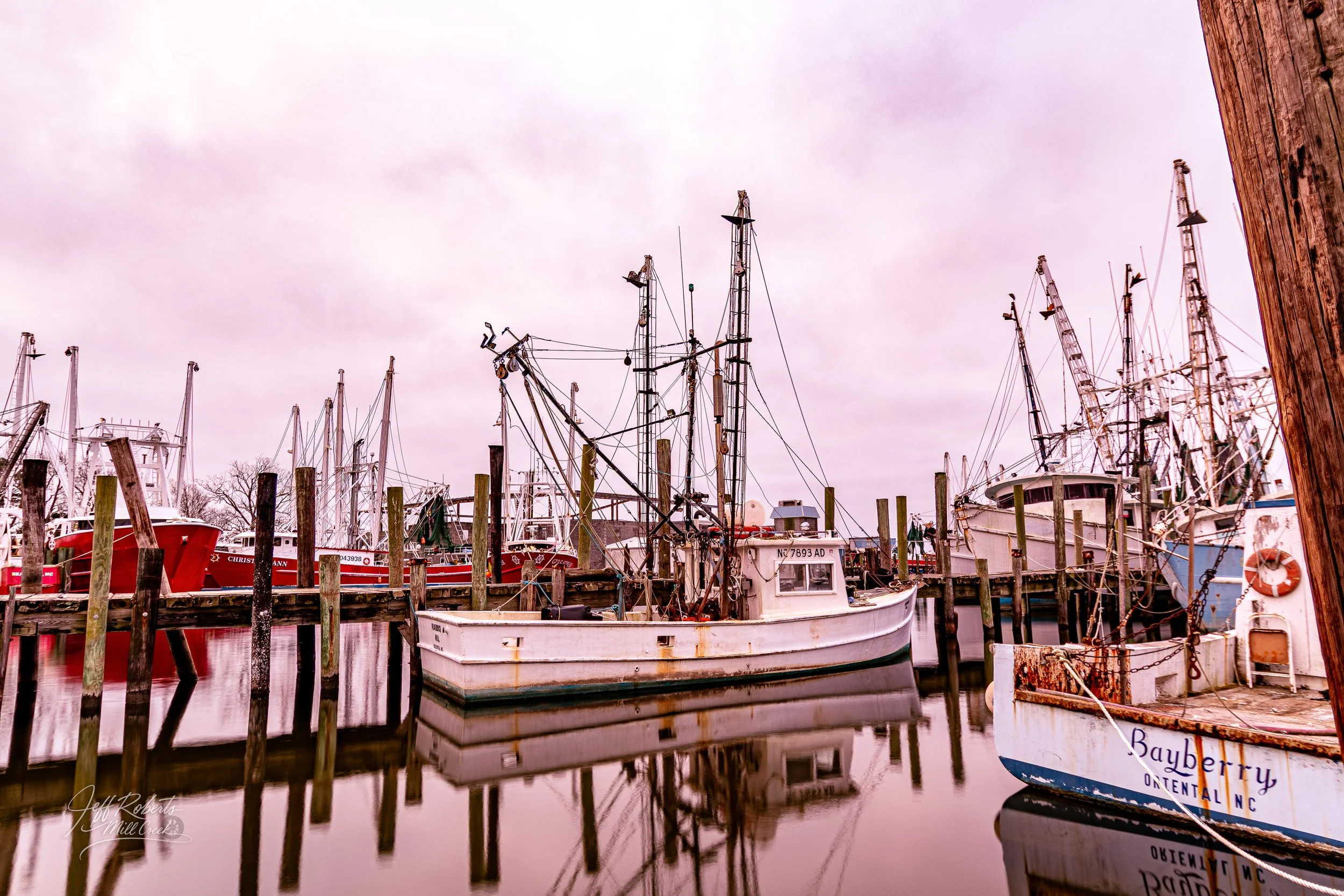 Fishing boats docked at a marina, with wooden posts and calm water reflecting the boats, under an overcast sky.