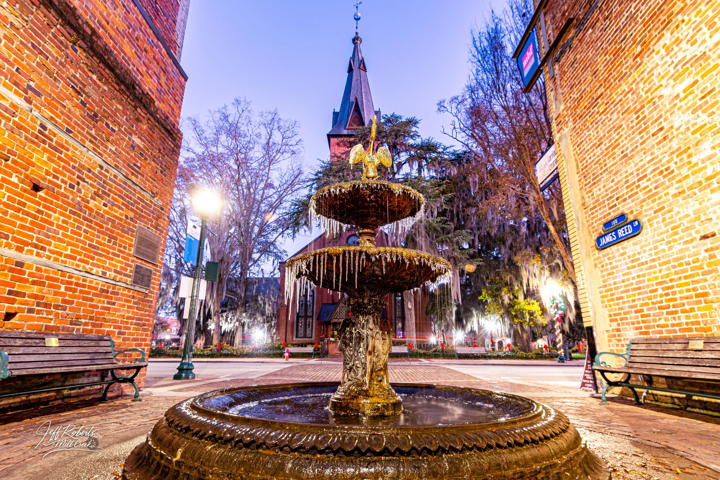 A historic brick building with a pointed steeple and a decorative fountain in front, decorated with Christmas ornaments, with benches and street signs visible.
