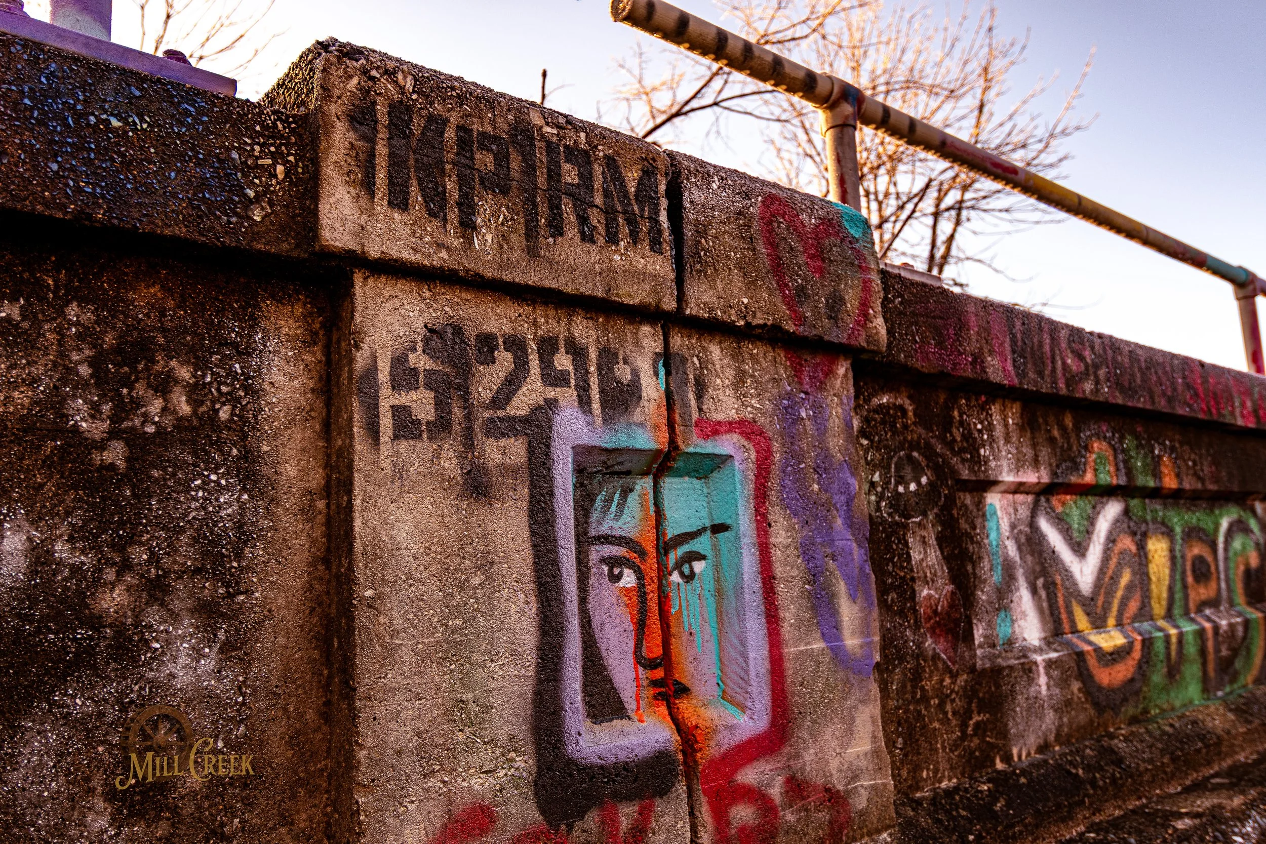 Concrete structure with colorful graffiti including a stylized face with eyes and nose, surrounded by various tags and numbers, with a metal pipe running overhead and leafless trees in the background.