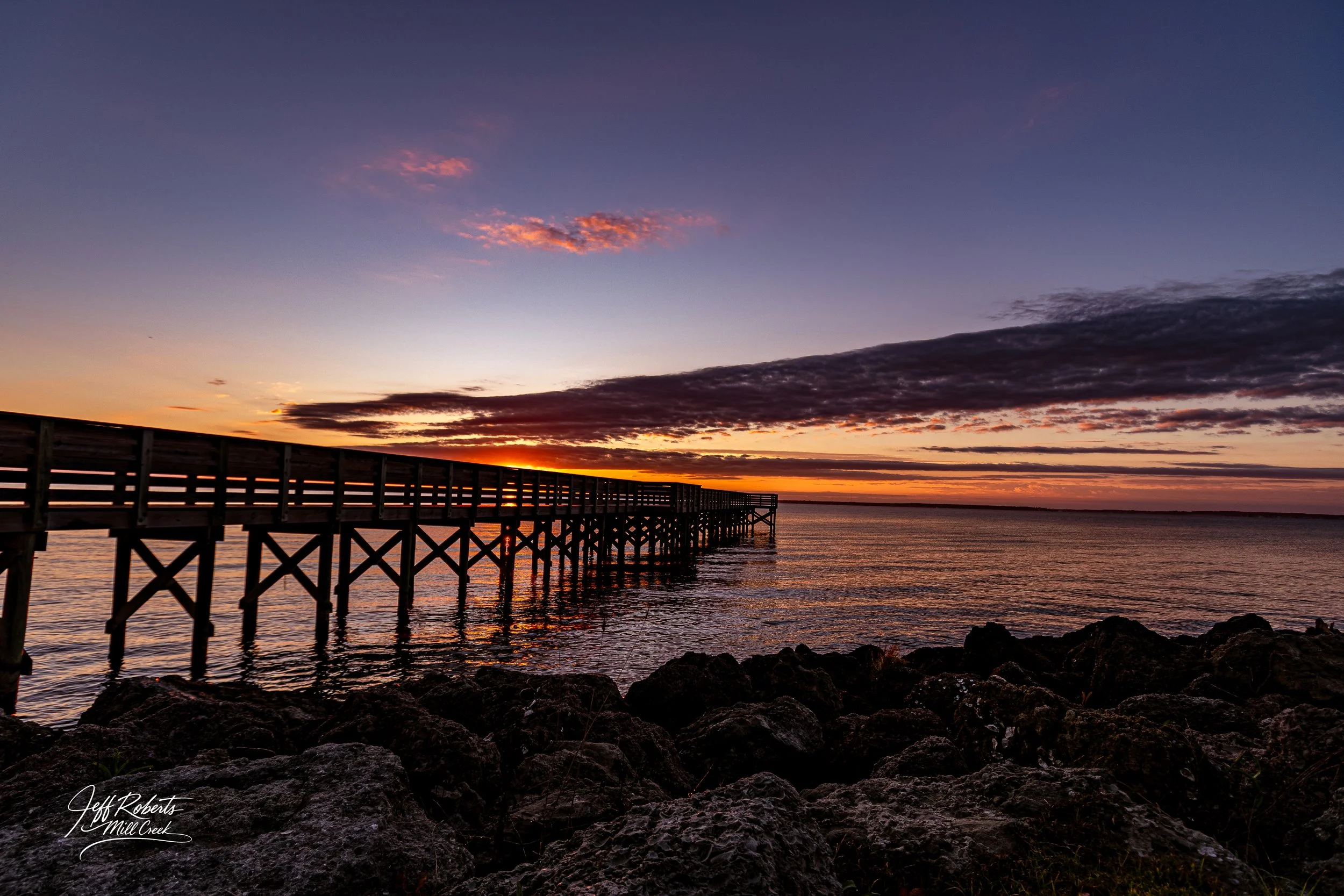 Sunset over a wooden pier extending over water with rocks in the foreground and clouds in the sky.