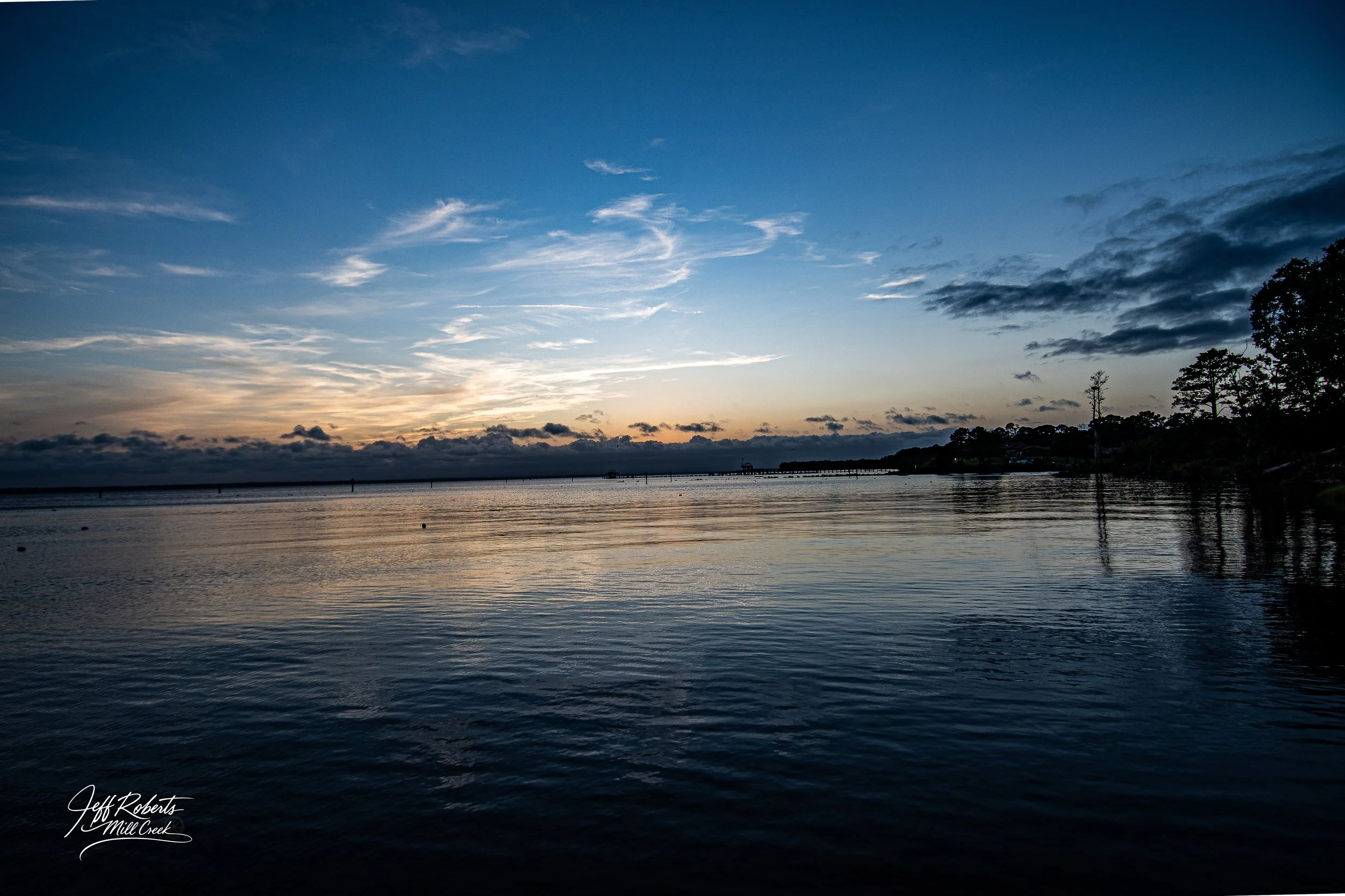 A serene river scene at sunset with a partly cloudy sky, calm water with gentle ripples, and trees along the shoreline in the distance.