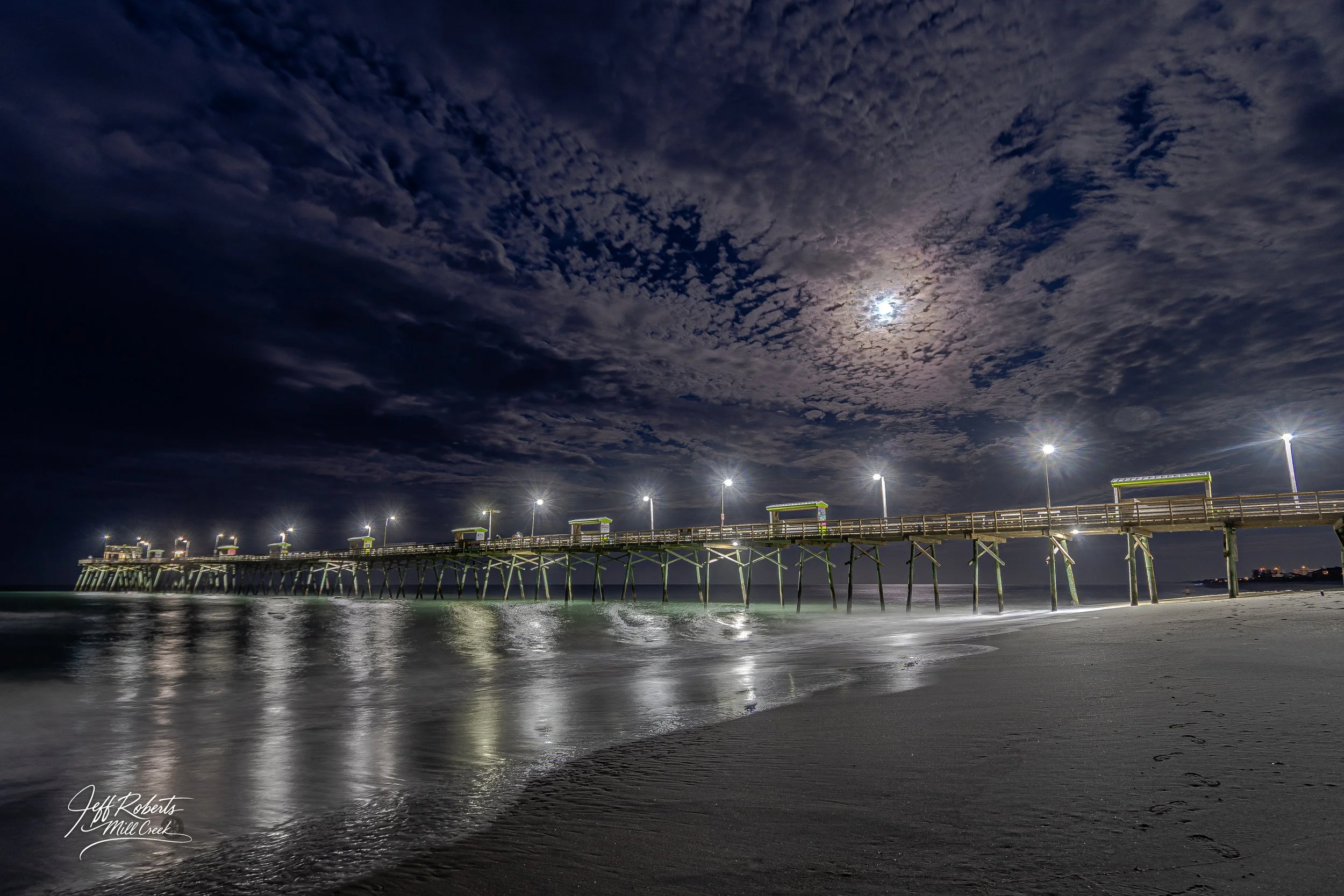 Night scene of a pier stretching over calm ocean waters under a cloudy sky with the moon partially visible and streetlights illuminating the pier.