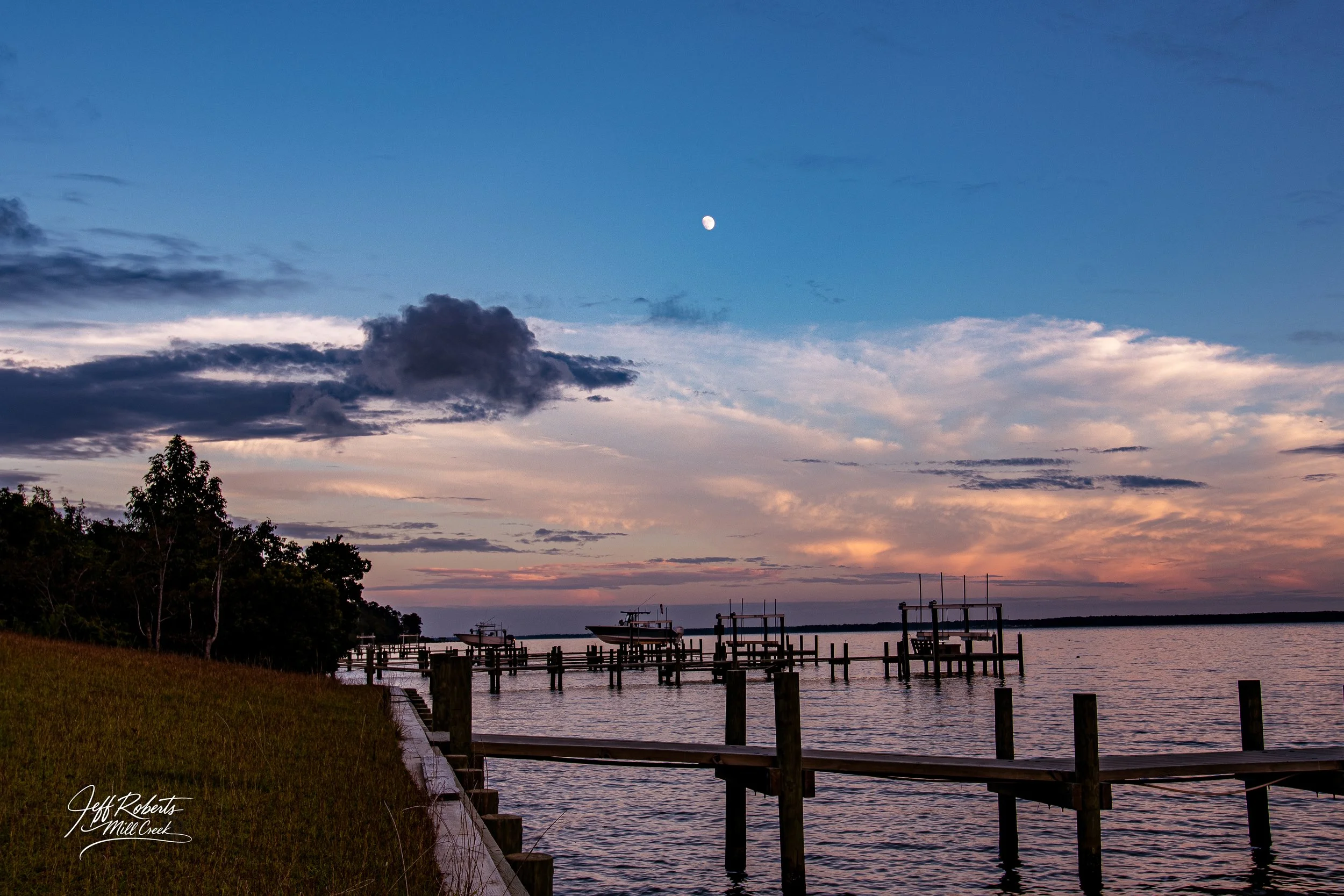 A tranquil body of water at sunset with a partly cloudy sky and a visible moon, a grassy shoreline on the left, trees, wooden docks extending into the water, and boats docked.