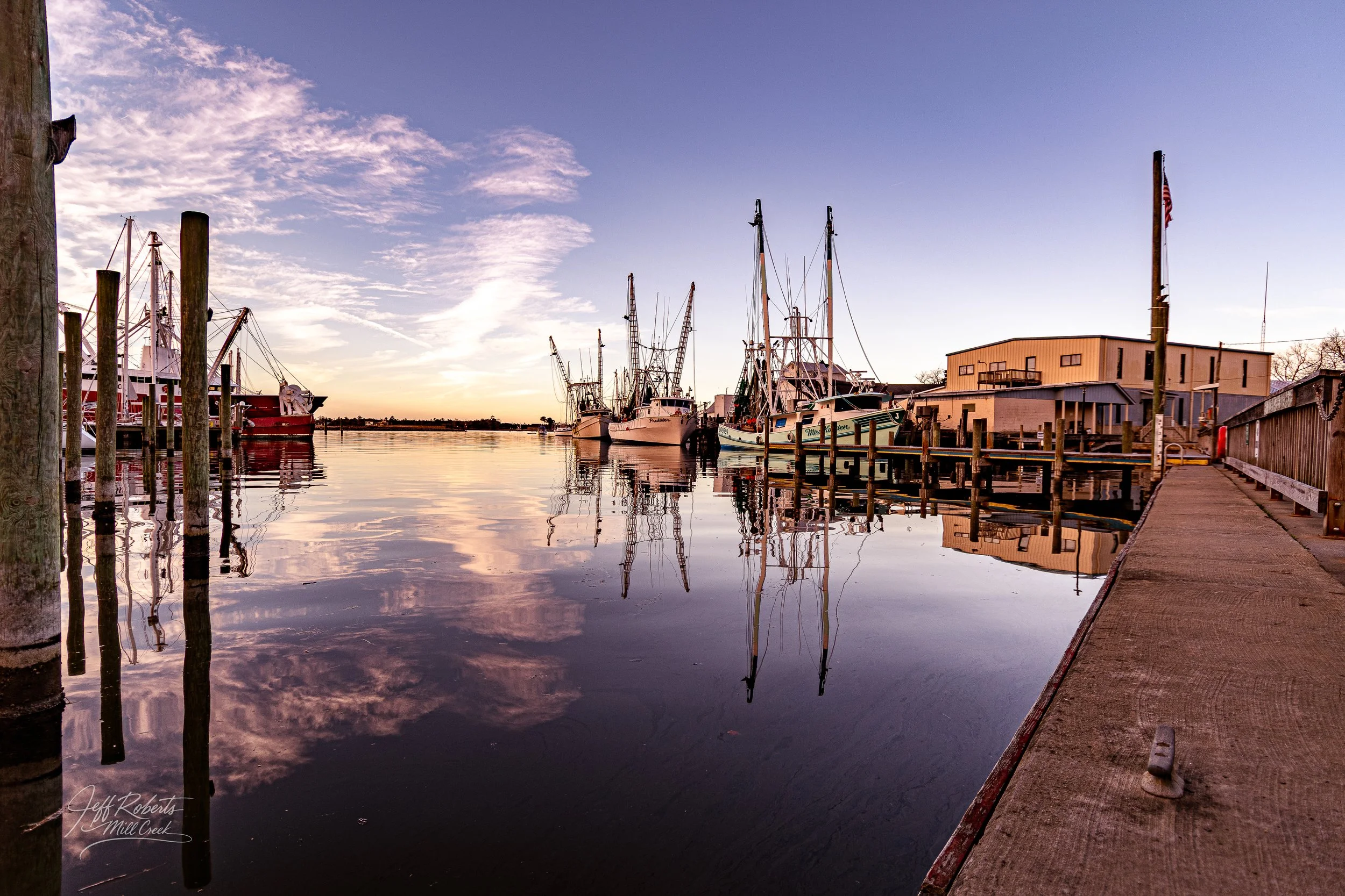 A peaceful marina scene at sunset with boats docked along the pier, reflecting on calm water, under a partly cloudy sky.