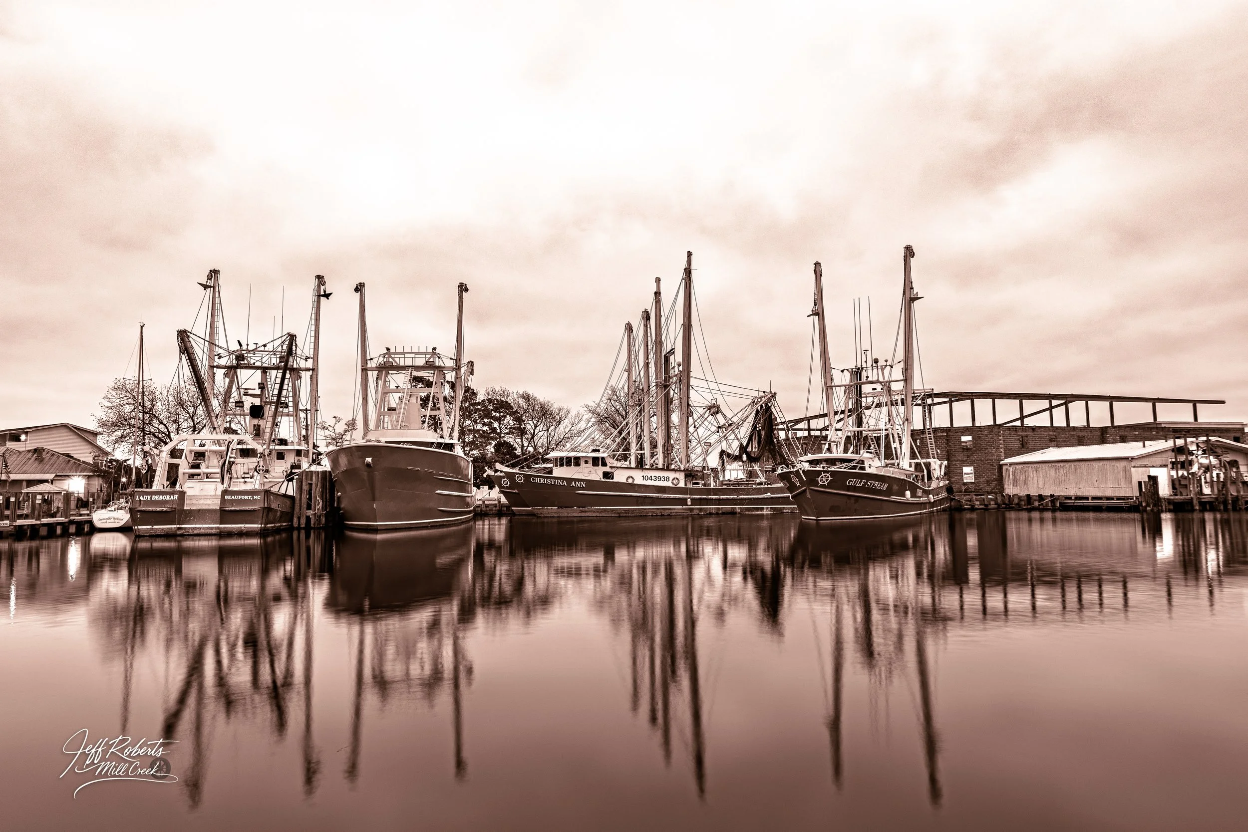 Boats docked at a marina with their reflections visible in calm water, under cloudy sky.