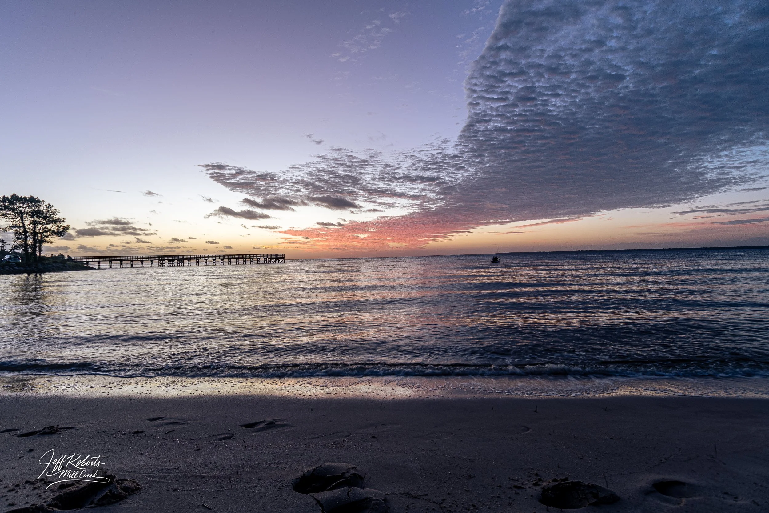 Sunset over the ocean with a partly cloudy sky, a wooden pier extending into the water, a small boat in the distance, and a sandy beach in the foreground.