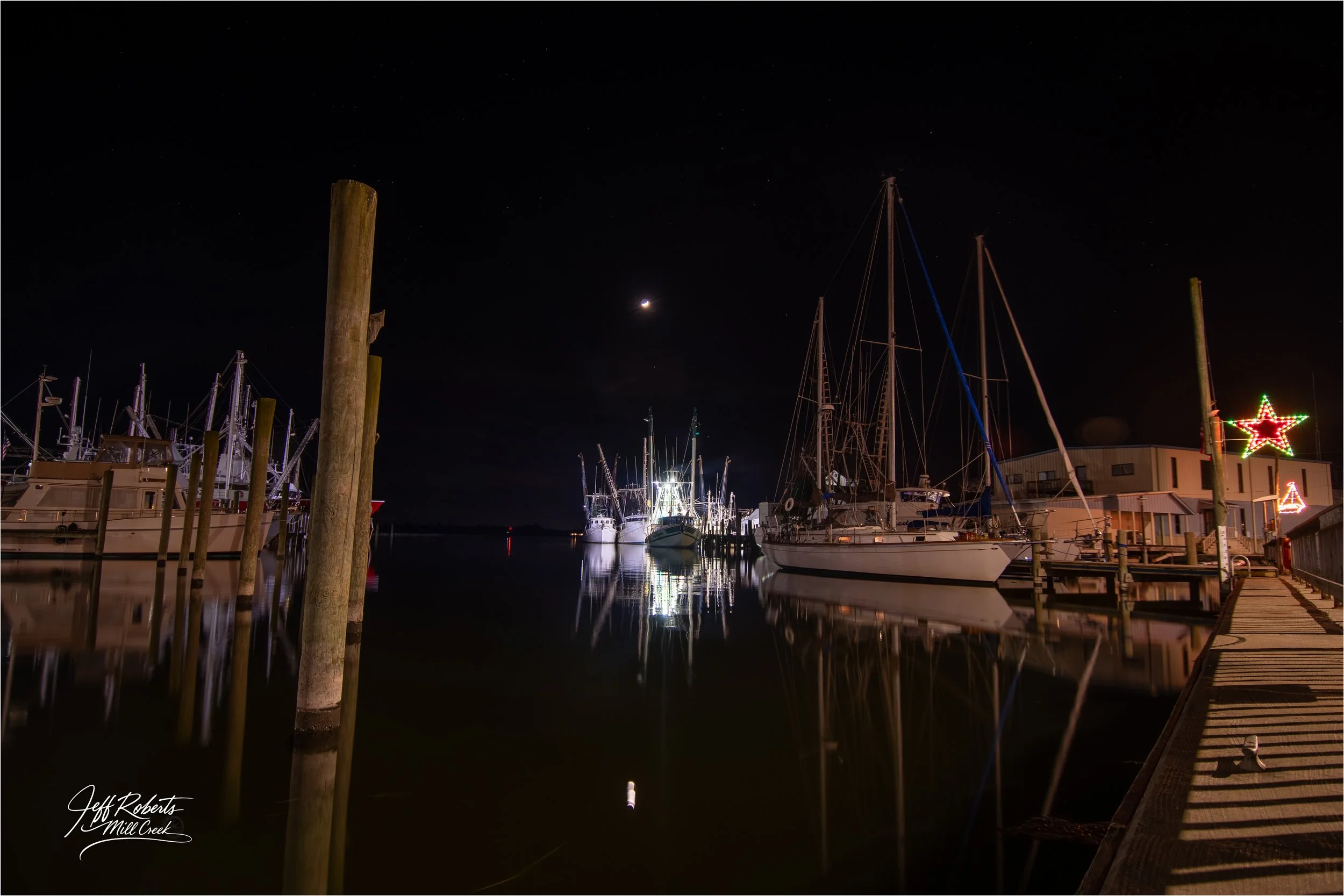 Nighttime view of a marina with docked boats, illuminated by artificial lights, under a moonlit sky with a few stars, and festive holiday decorations on a nearby building.