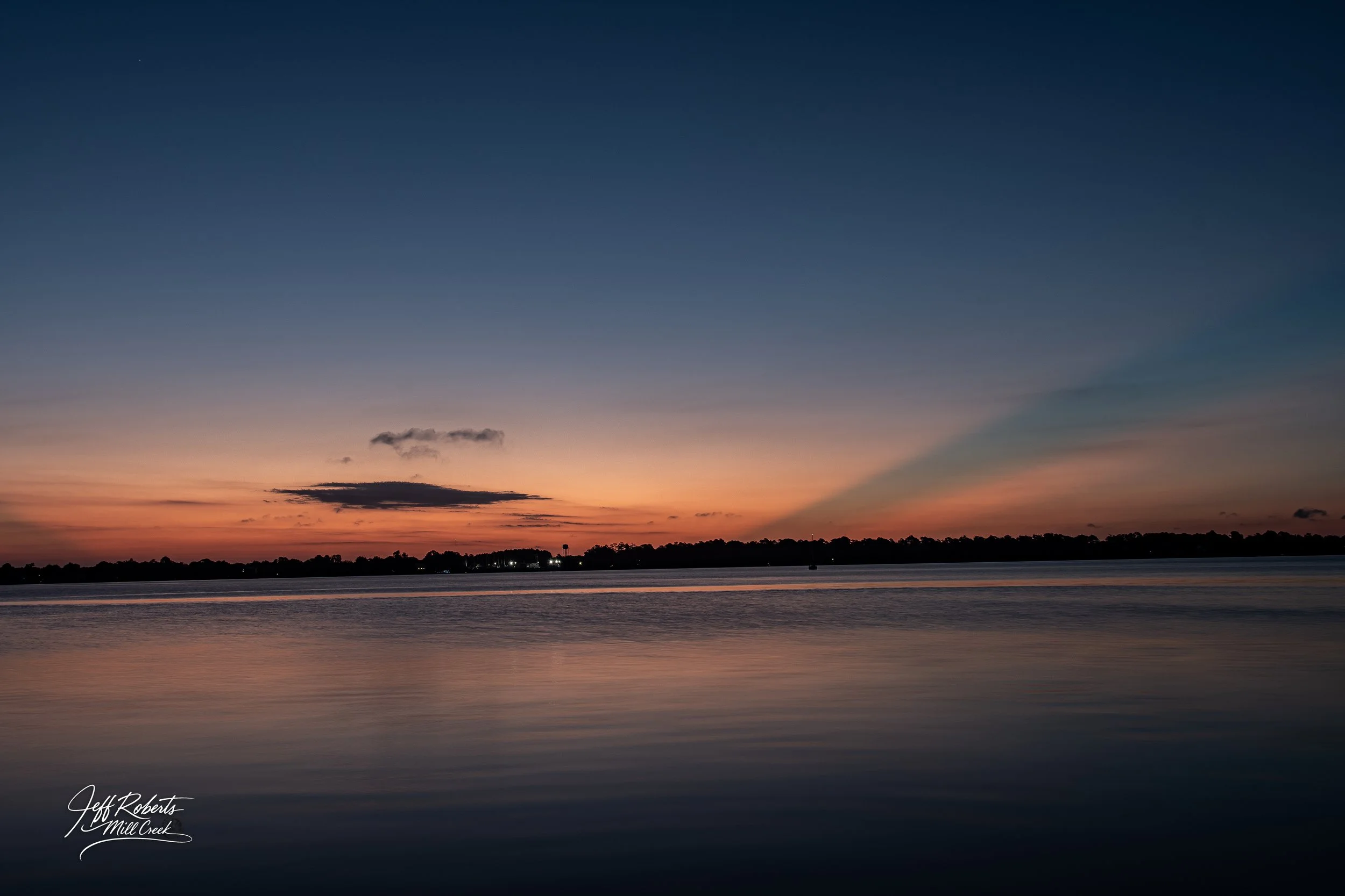 Sunset over a calm river with a silhouette of trees on the horizon and a few clouds in the sky.