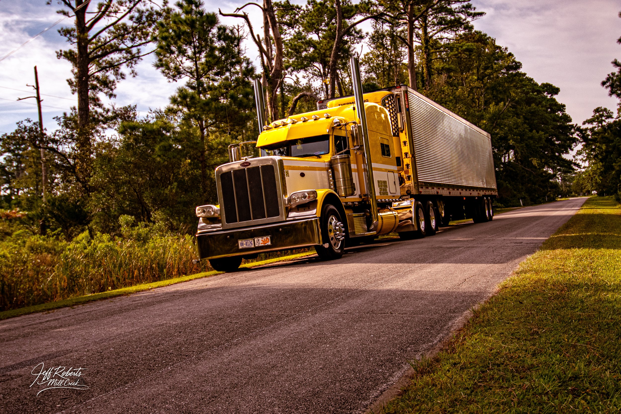 A yellow semi-truck with a large grille and silver trailer driving on a rural road surrounded by trees and grass.
