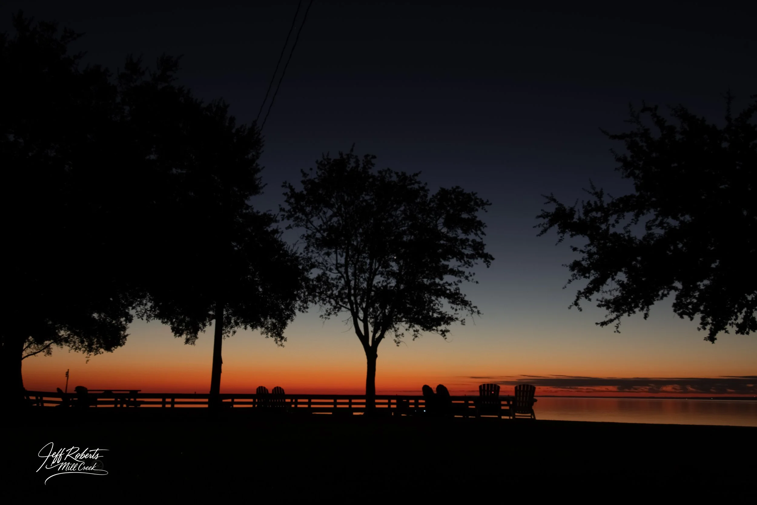 Silhouettes of trees and benches along a lakeside at sunset with orange, pink, and blue sky reflections on the water.