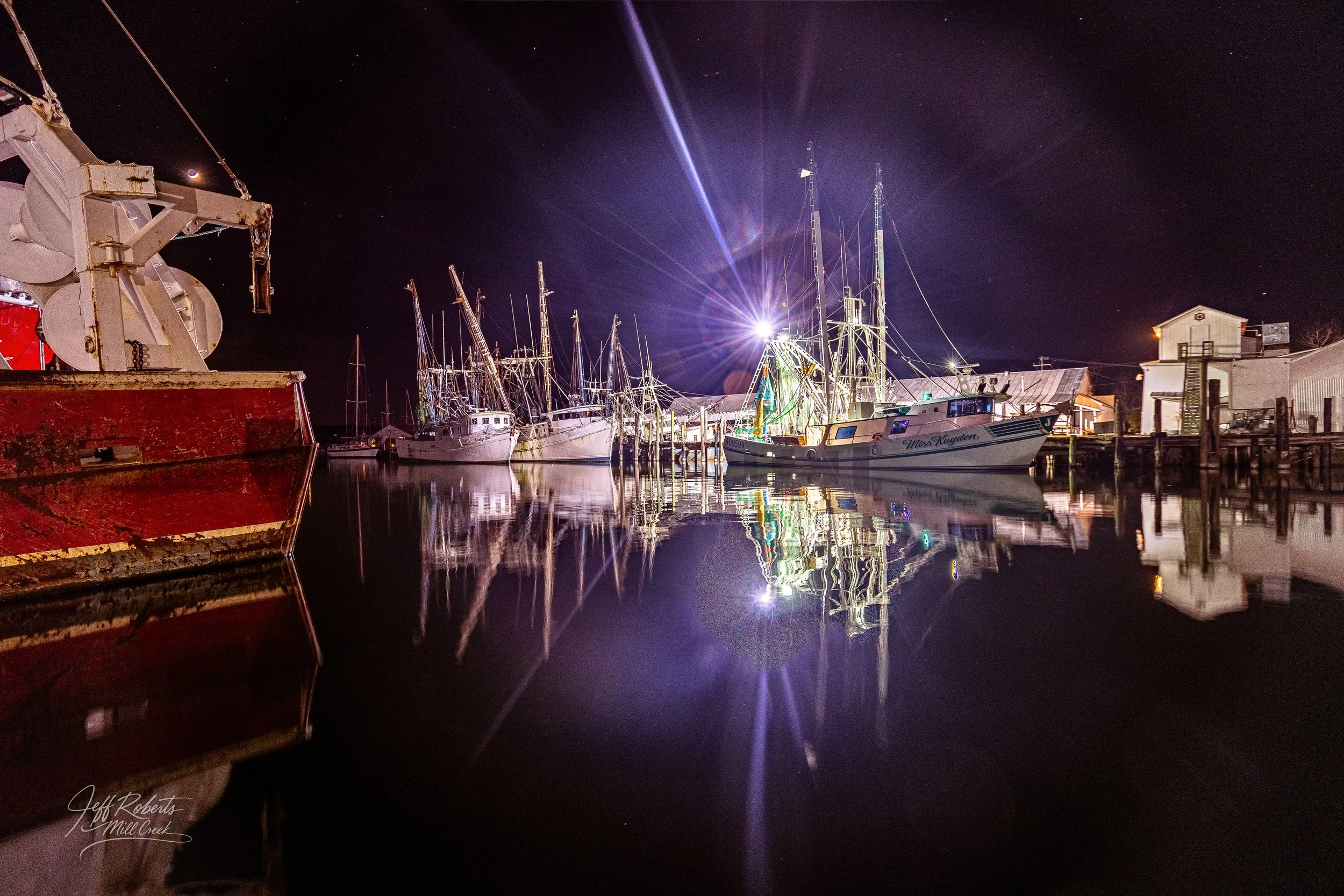 Nighttime view of boats docked at a marina, with reflections on calm water and a bright light shining on the boats