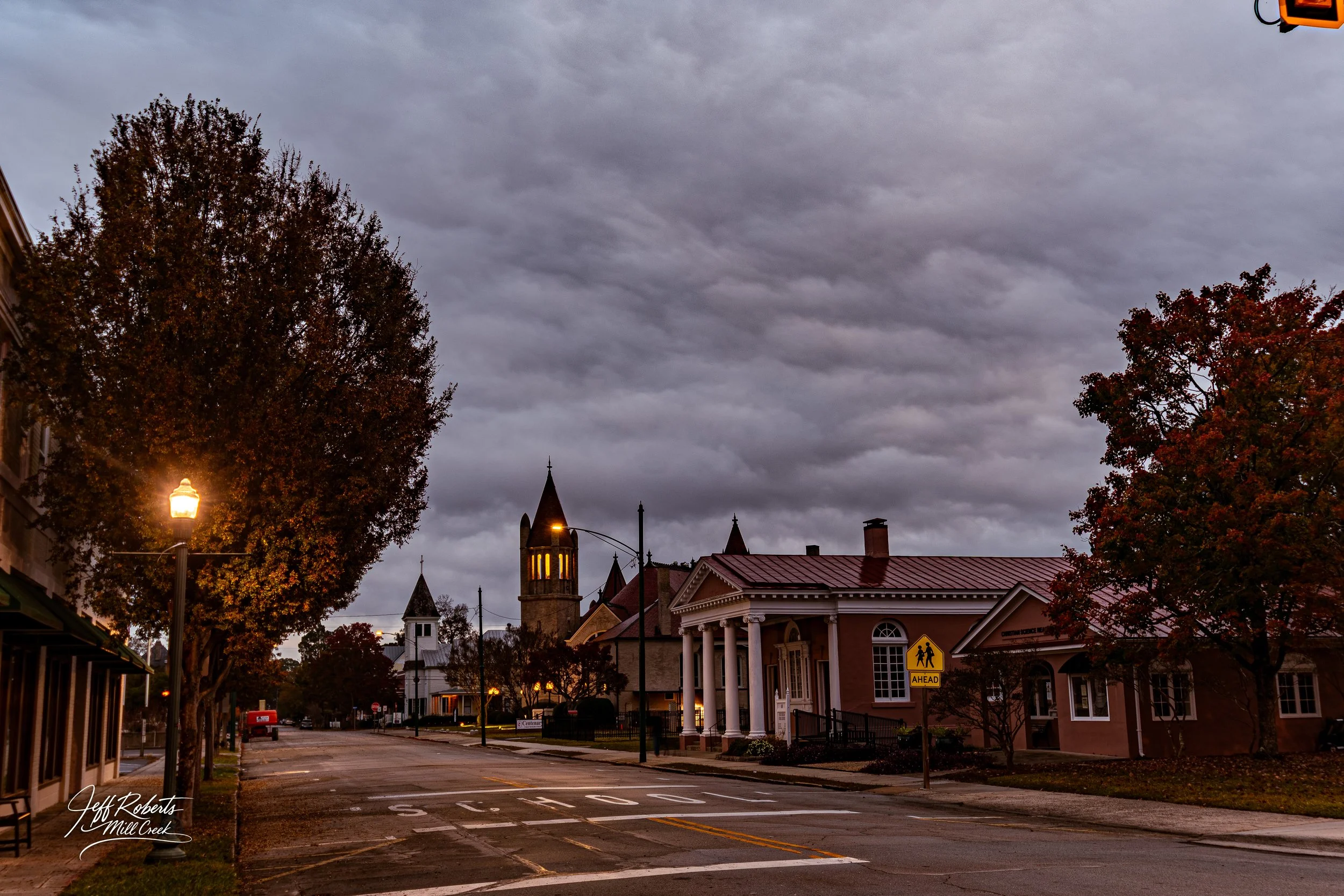 Empty street in a small town during dusk with cloudy skies, with trees, a church with a tall steeple, and a building with columns, streetlights on, and a yellow school zone sign.