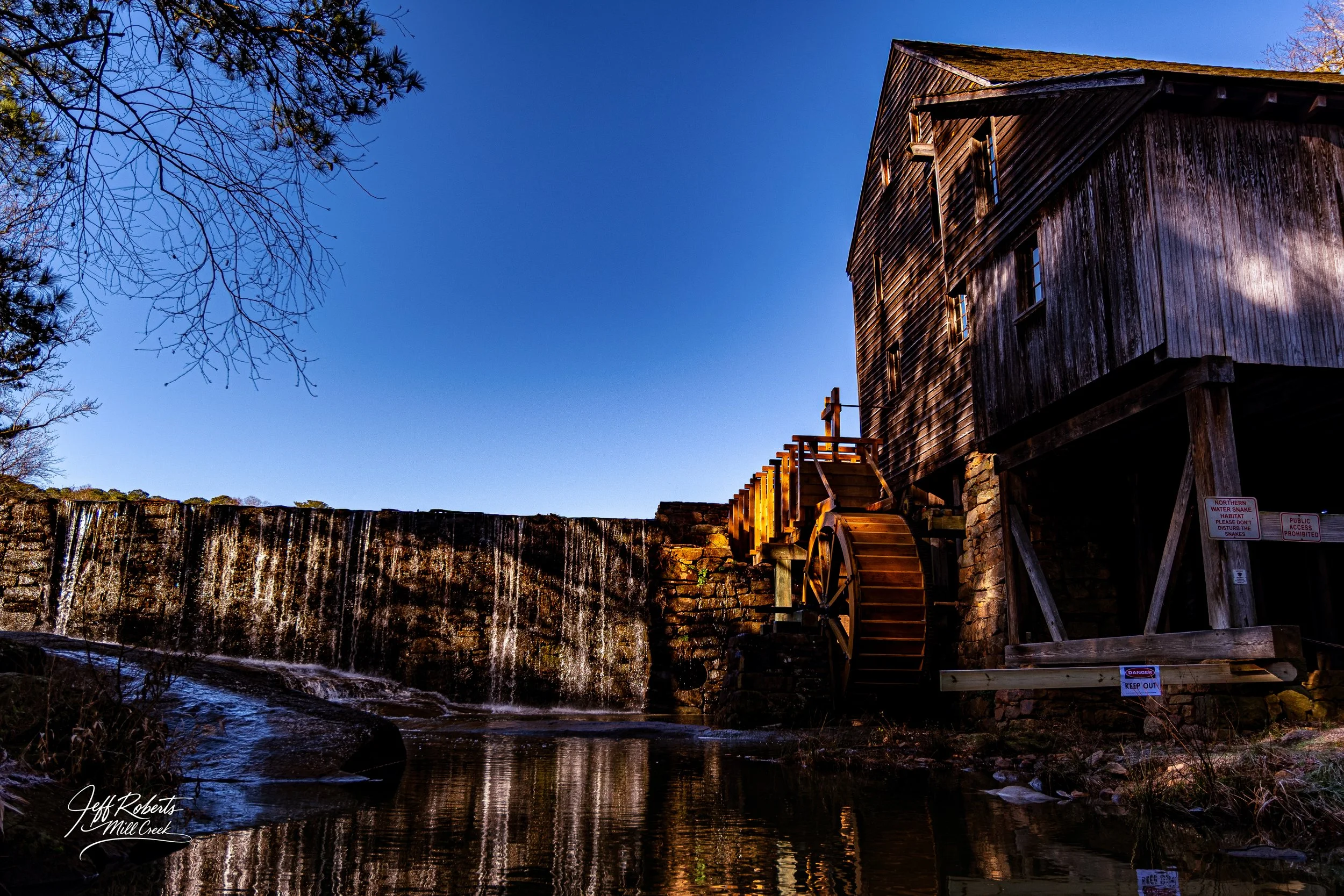 Old wooden watermill beside Mill Creek with a small waterfall and a clear blue sky.
