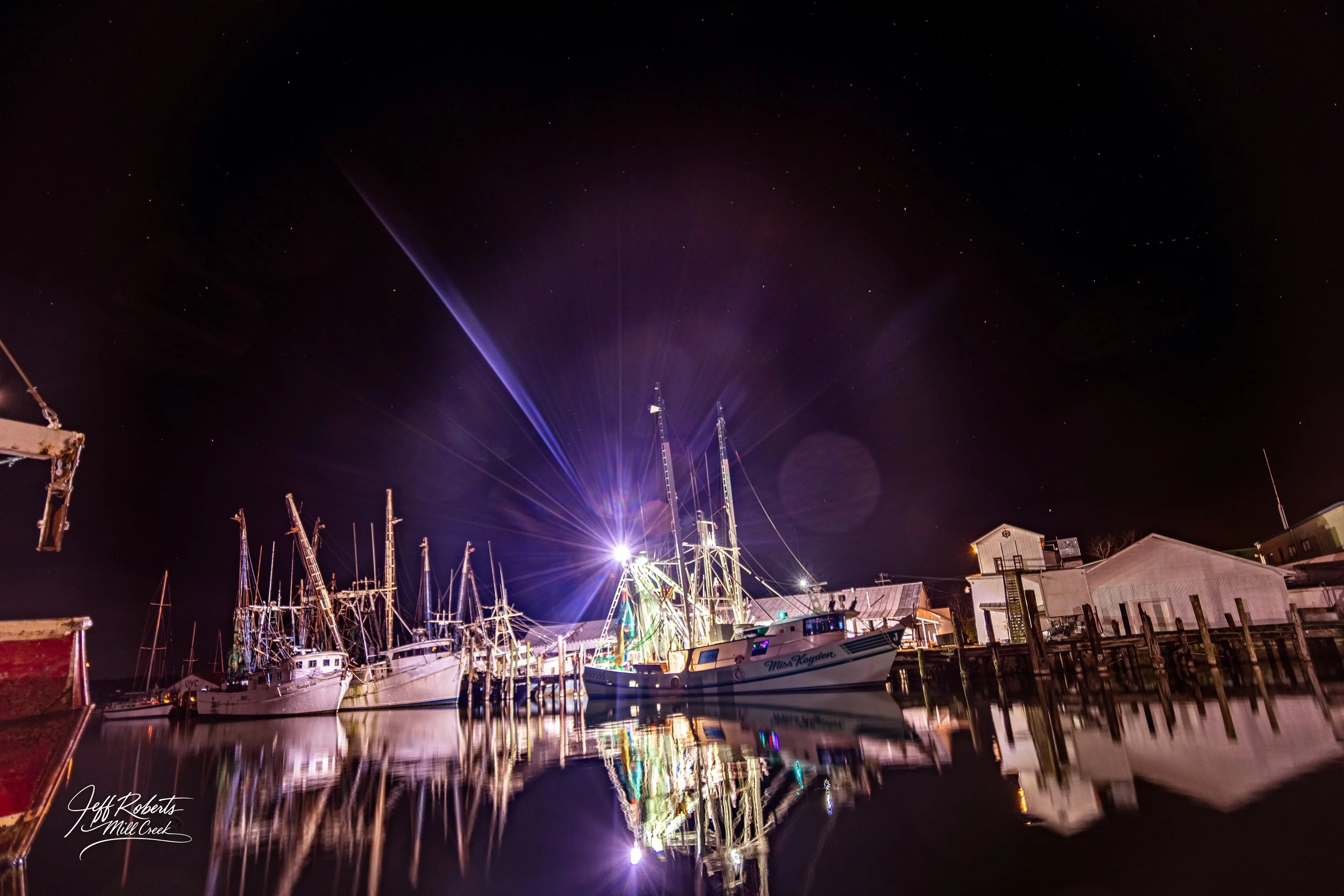 A nighttime scene at a marina with boats docked at the pier, illuminated by bright artificial lights, with their reflections visible on the still water surface and a starry sky overhead.