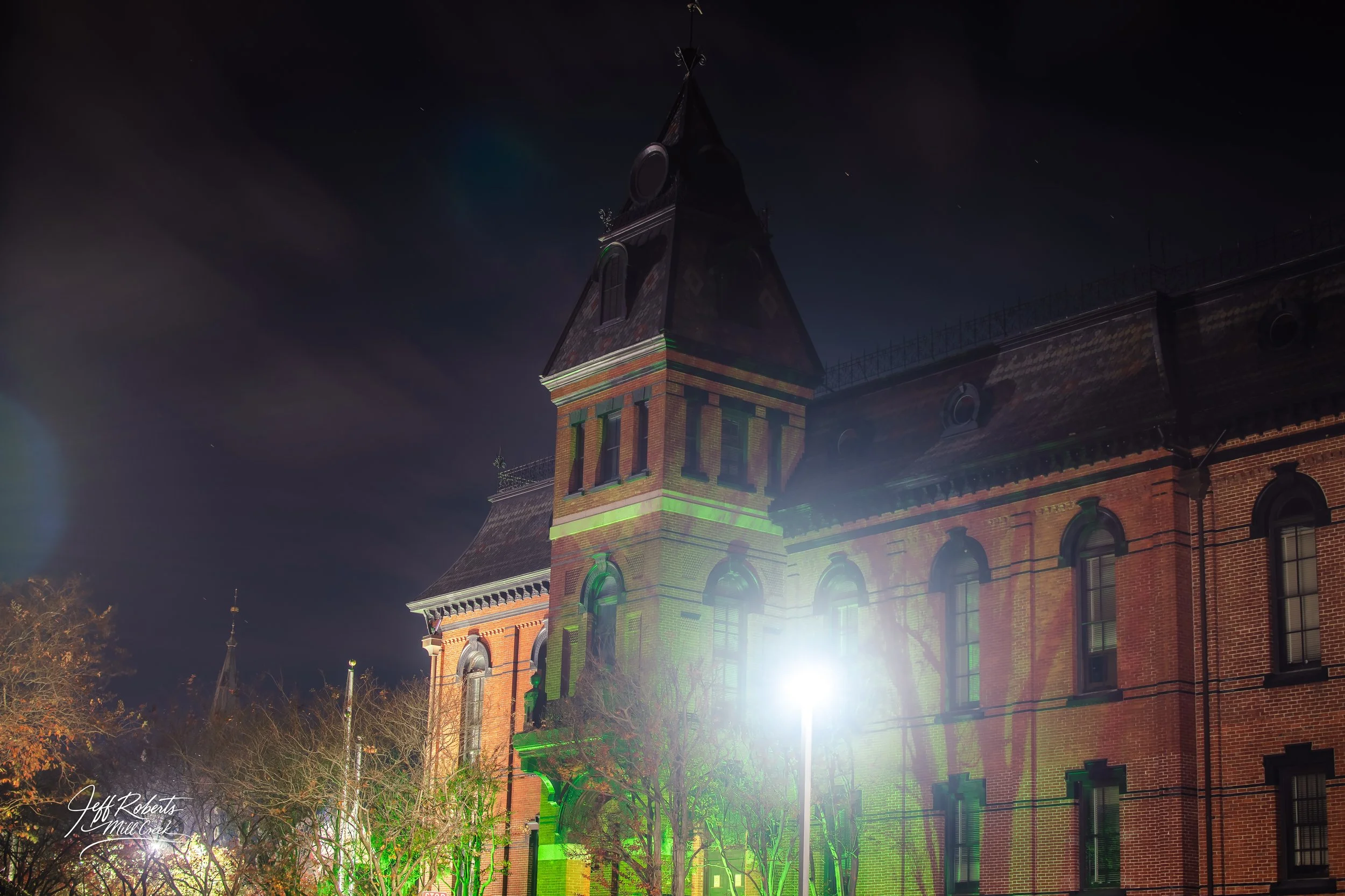Night view of a historic brick building with a tower, lit by streetlights, with trees in front and a dark sky overhead.