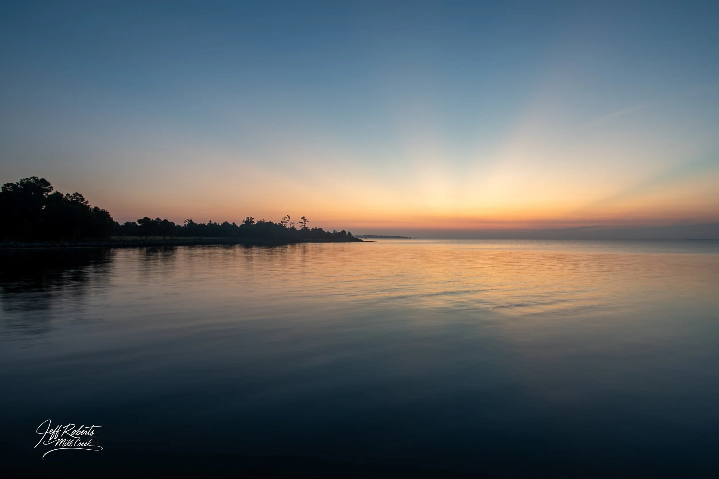 Sunset over a calm body of water with a tree-lined shoreline on the left and a colorful sky with gradient from blue to orange.