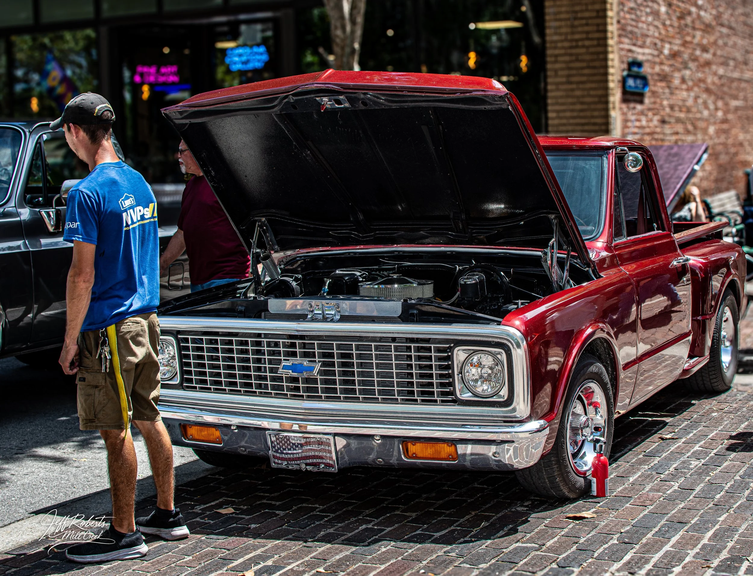 Red vintage Chevrolet pickup truck with open hood parked on a cobblestone street during a car show, with a fire extinguisher near the front tire and two people standing nearby.