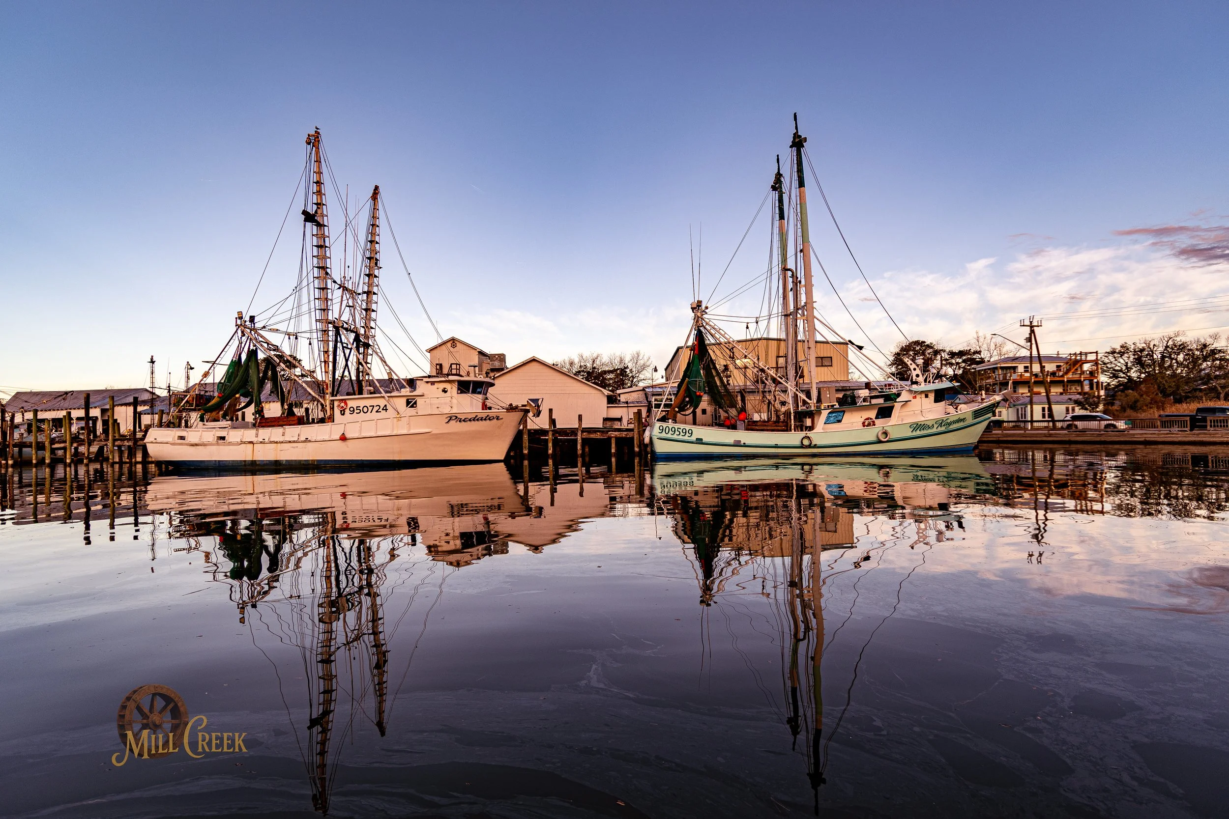 Two boats docked at a marina with buildings and trees in the background during sunset, reflecting on calm water.