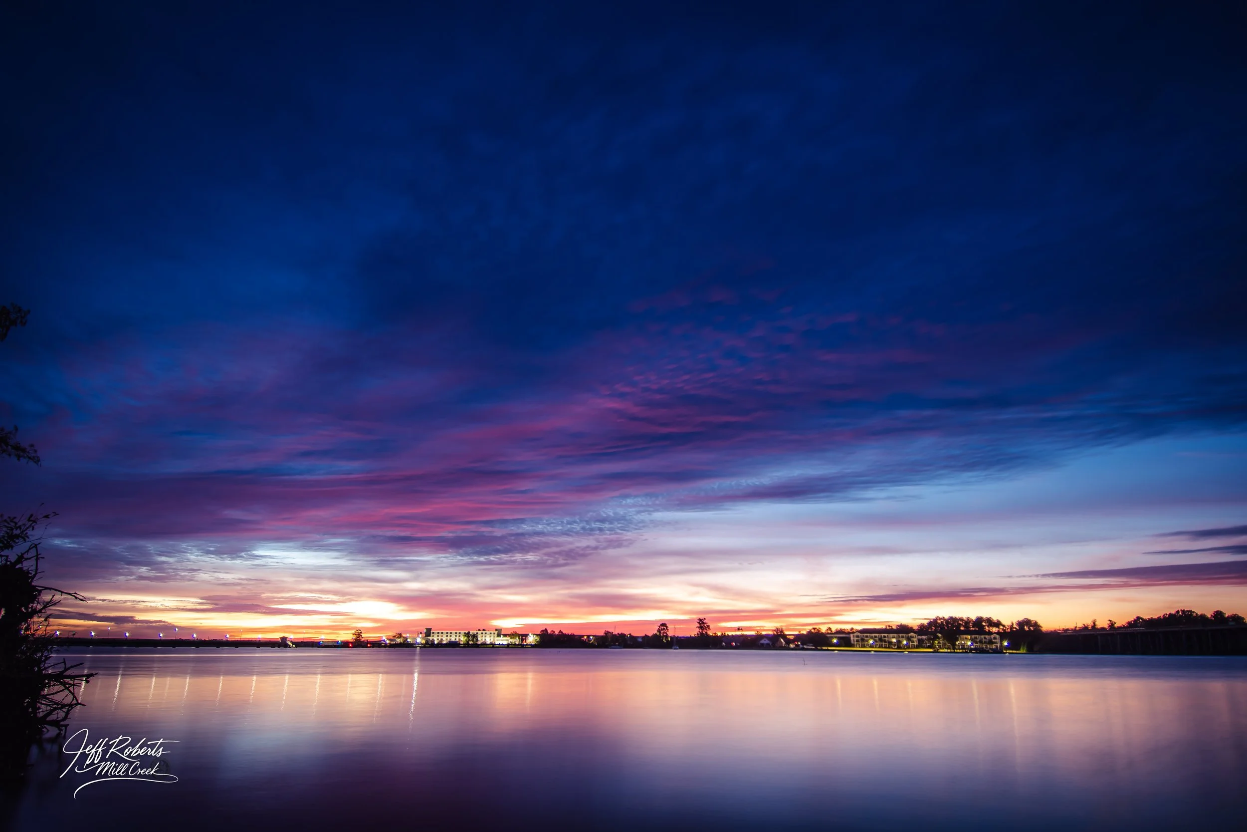 A colorful dusk sky with shades of blue, pink, and orange over a calm lake, with city lights and silhouettes of trees on the distant shoreline.