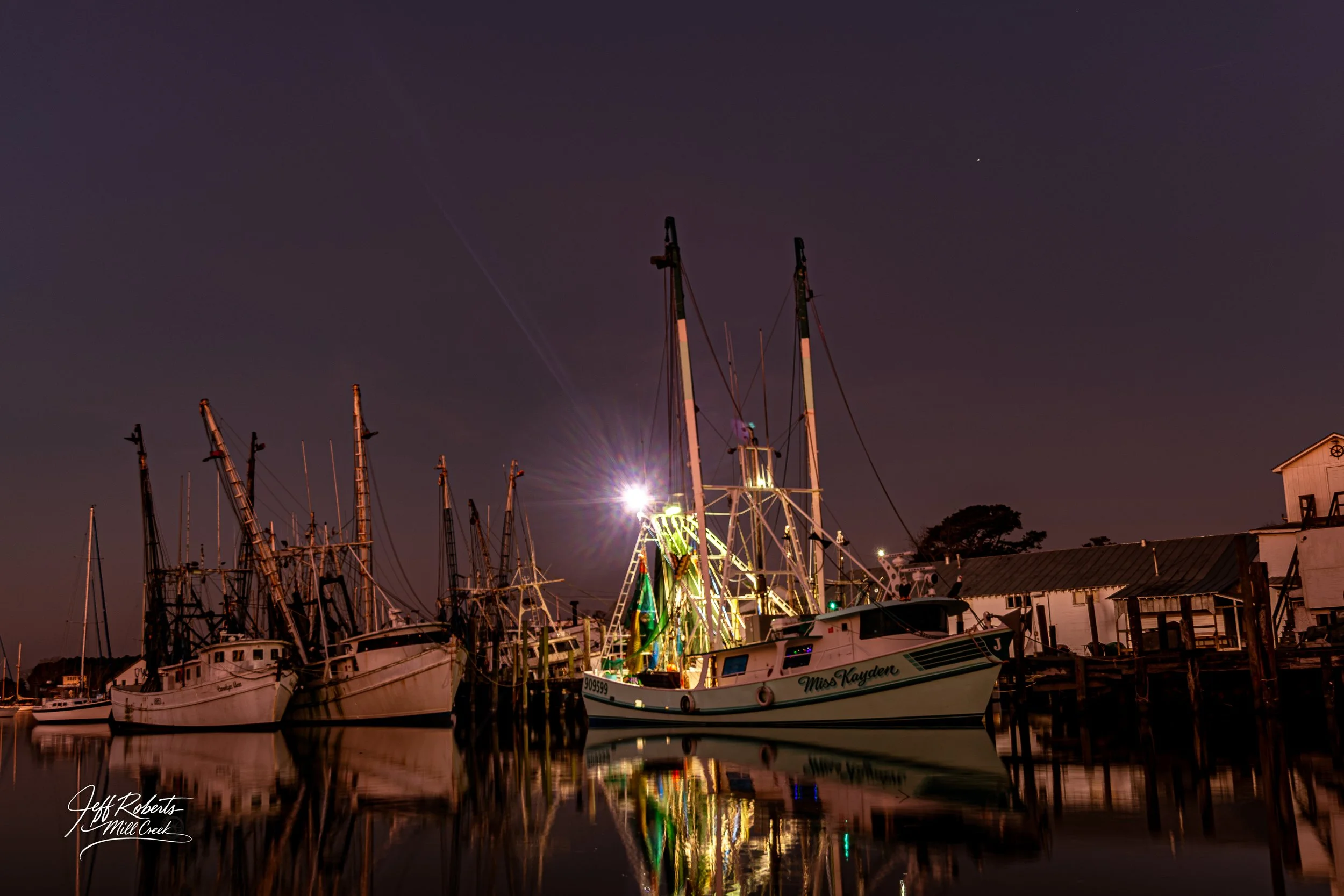 A marina at night with several docked boats and tall masts, illuminated by a bright light, with a calm water reflection, a building to the right, and a dark sky with a few stars.