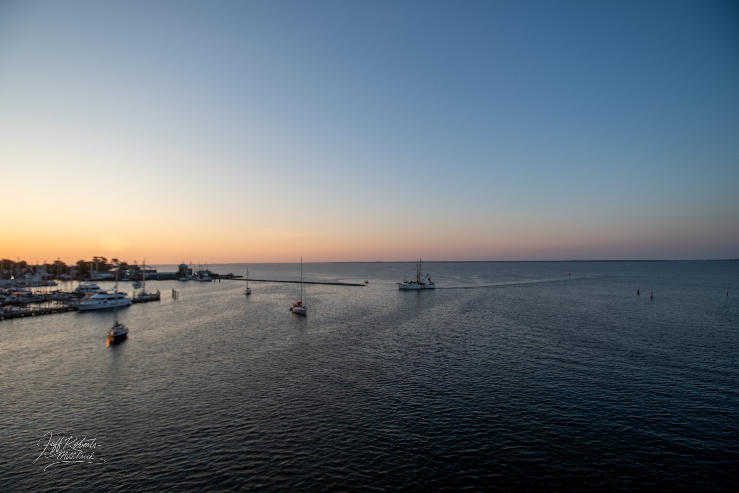 A harbor scene at sunset with sailboats anchored in calm water and a clear sky with a gradient from light orange near the horizon to blue at the top.