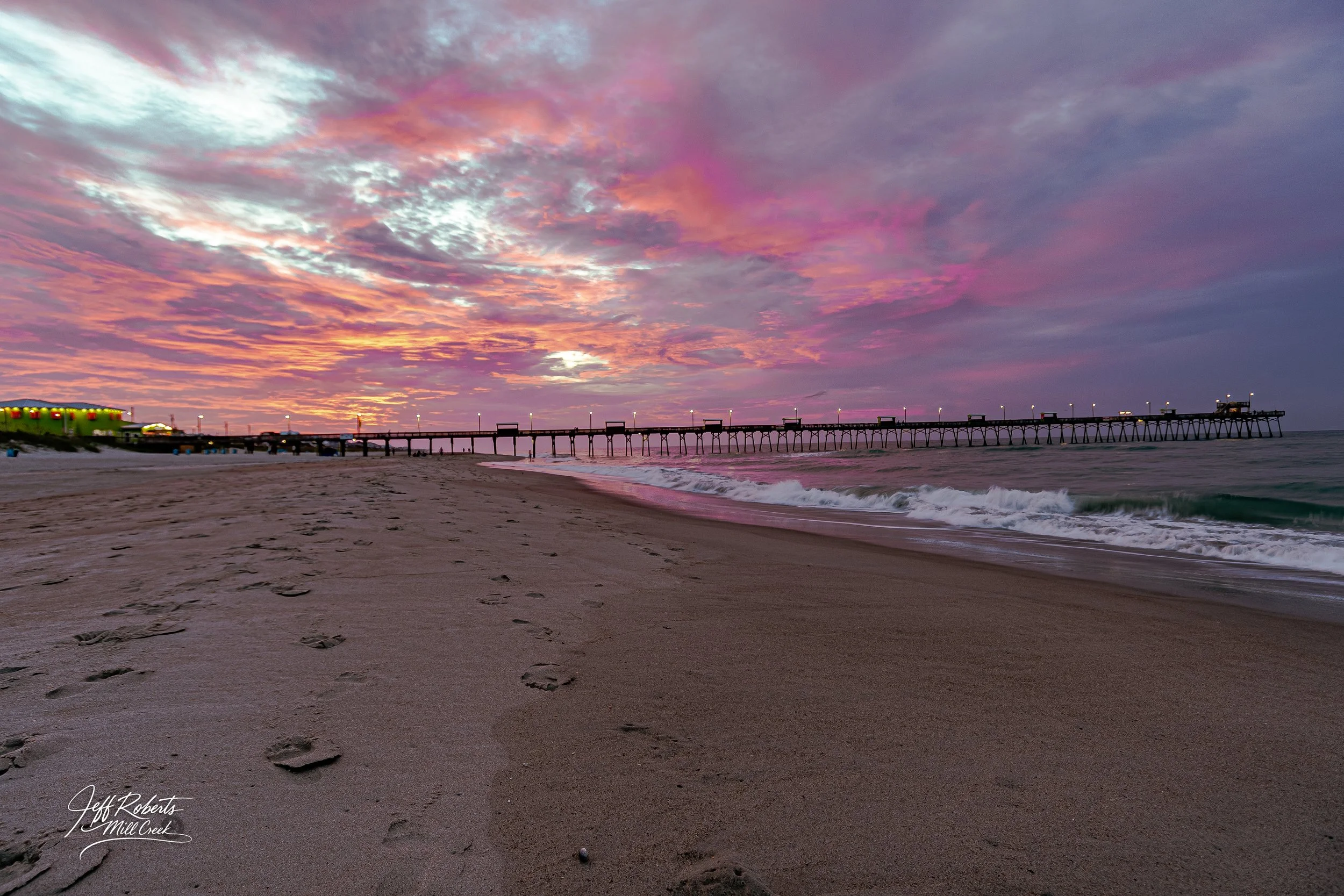 A sunset over a beach with footprints in the sand, a pier extending into the ocean, and colorful sky with pink, purple, and orange clouds.