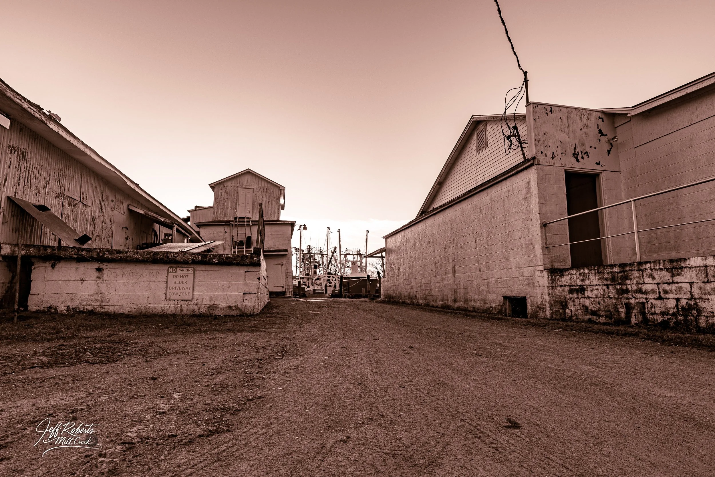 A dirt alleyway between old, weathered buildings with boats and a marina visible in the distance, under a pinkish sky.