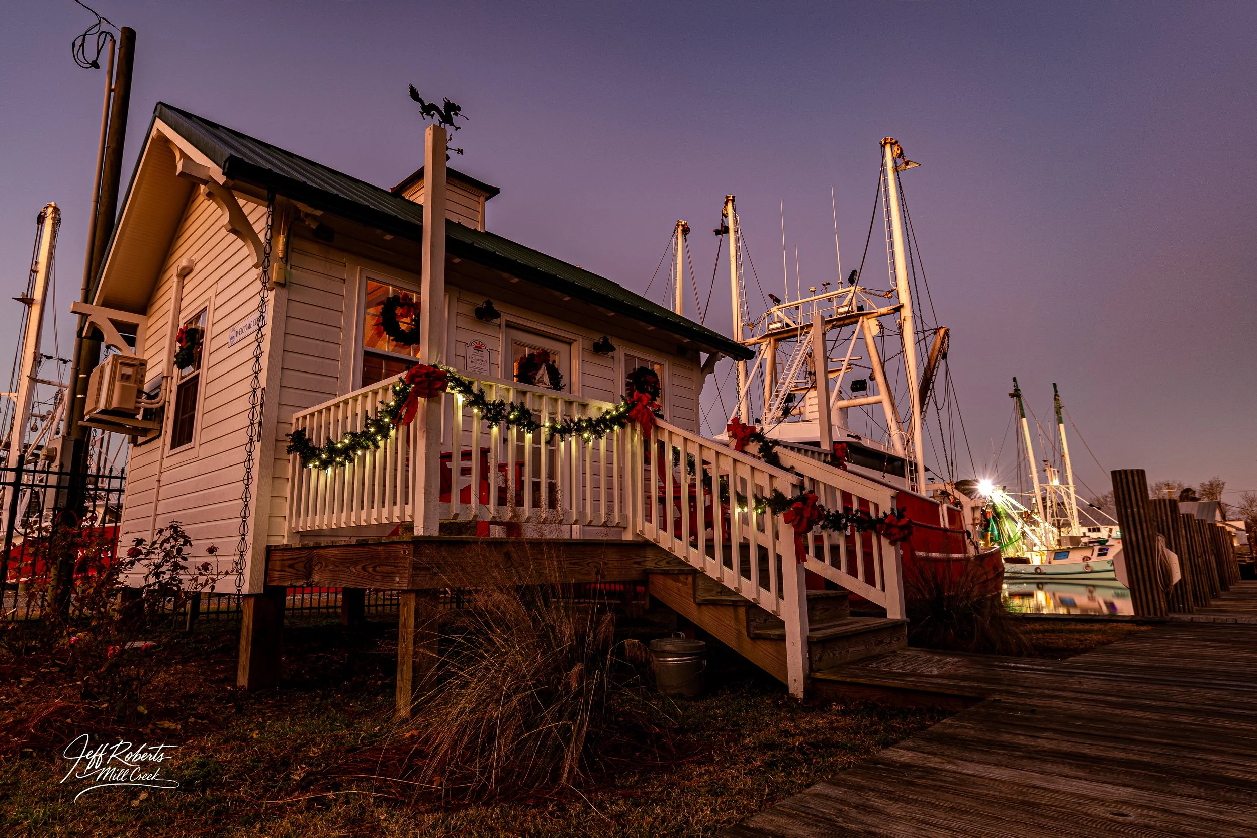 A festive house decorated for Christmas with garland and wreaths, situated next to a marina with boats, during twilight.
