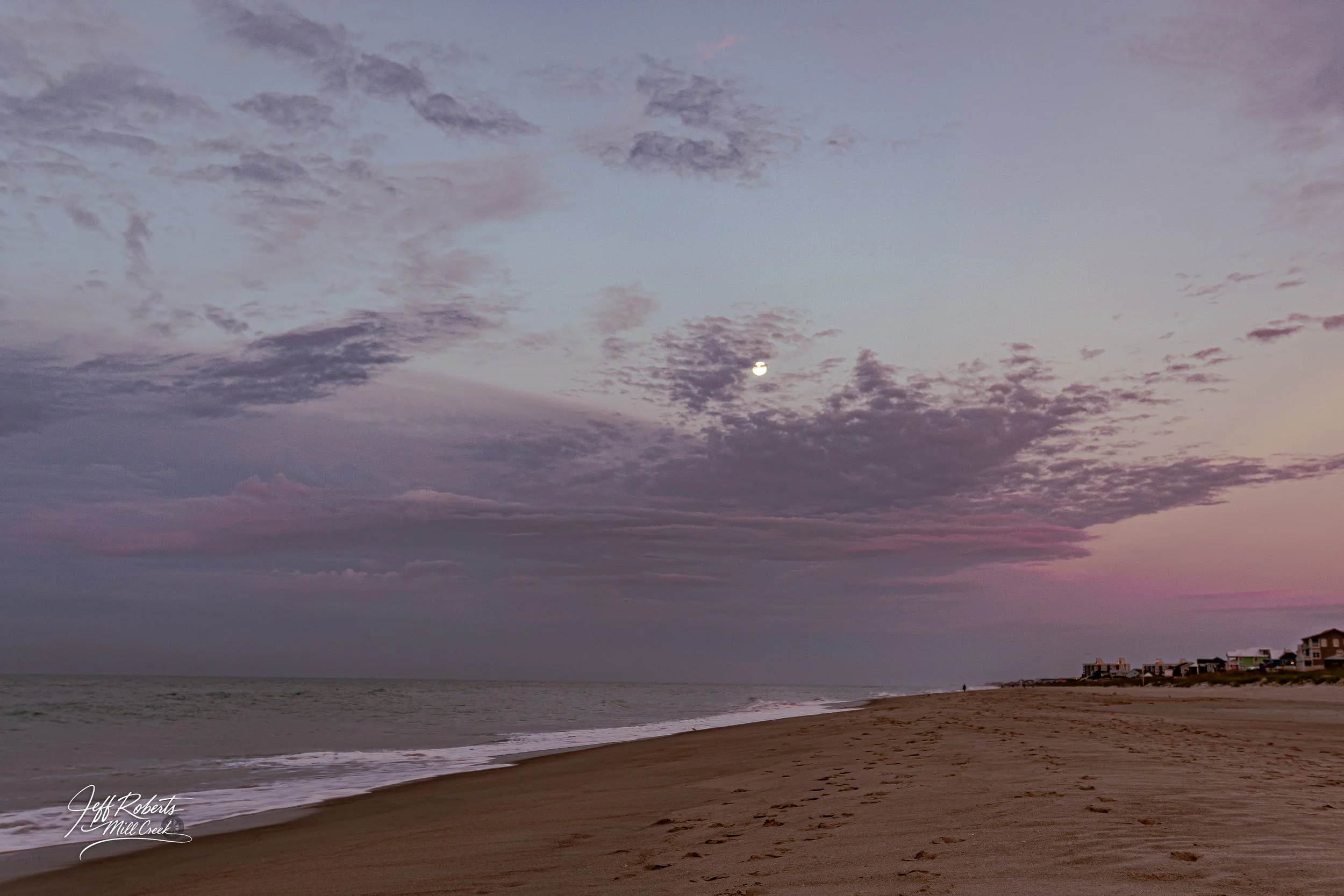A calm beach scene at dusk with footprints in the sand, a cloudy sky with the moon visible, and houses in the distance on the horizon.