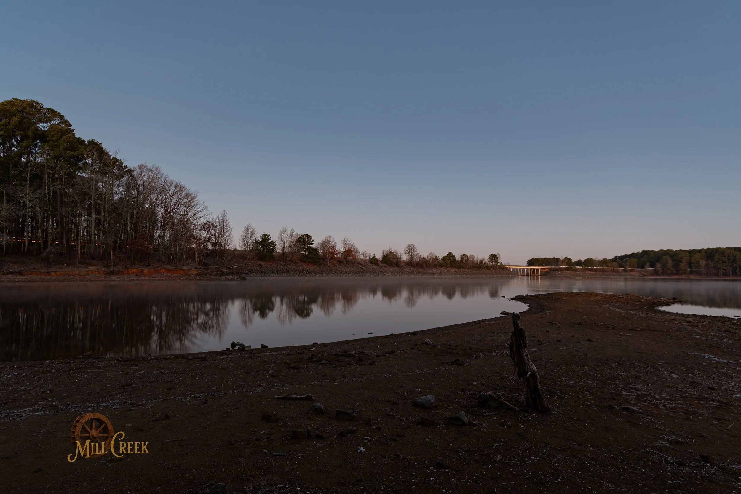 A calm body of water reflecting leafless trees along the shoreline, with a clear sky above and the Mill Creek logo in the bottom left corner.