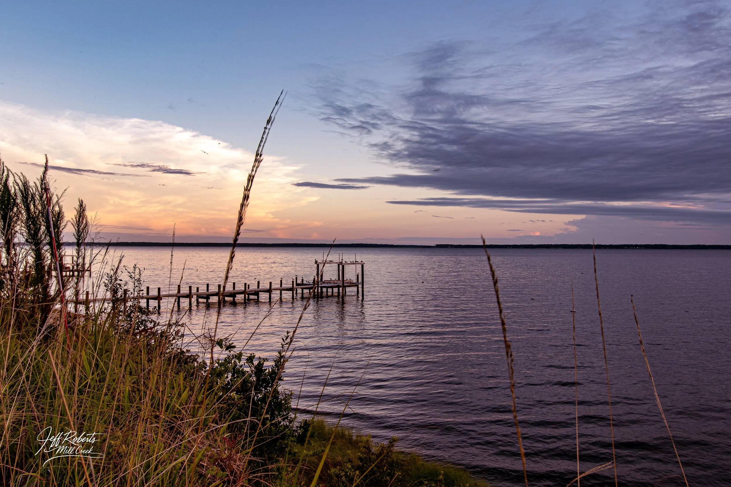 A lakeside scene at sunset with a wooden pier extending into calm water, tall grasses and bushes in the foreground, and a partly cloudy sky with hues of orange, pink, and purple.