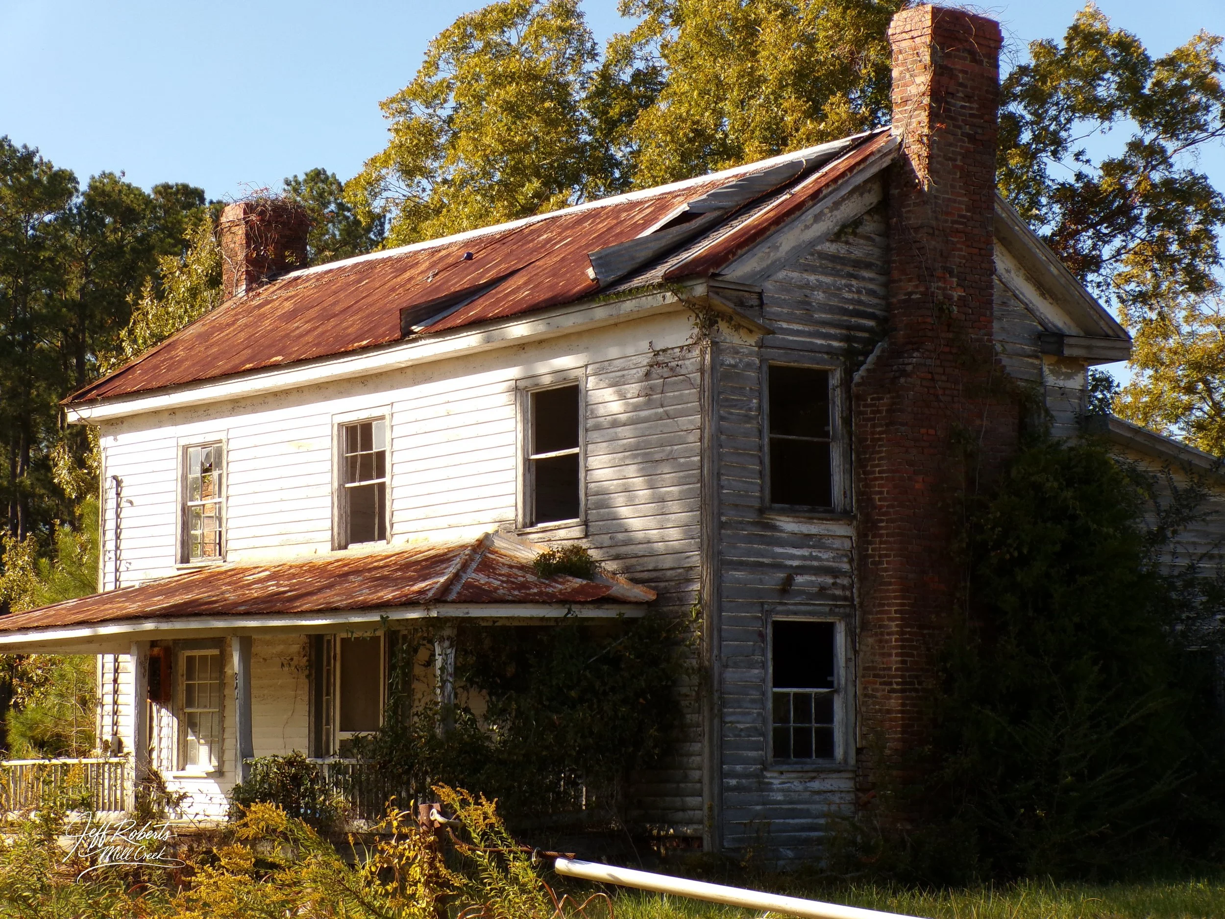An abandoned two-story house with weathered, peeling white siding, broken windows, rusted metal roof, and a tall brick chimney, surrounded by overgrown vegetation and trees.