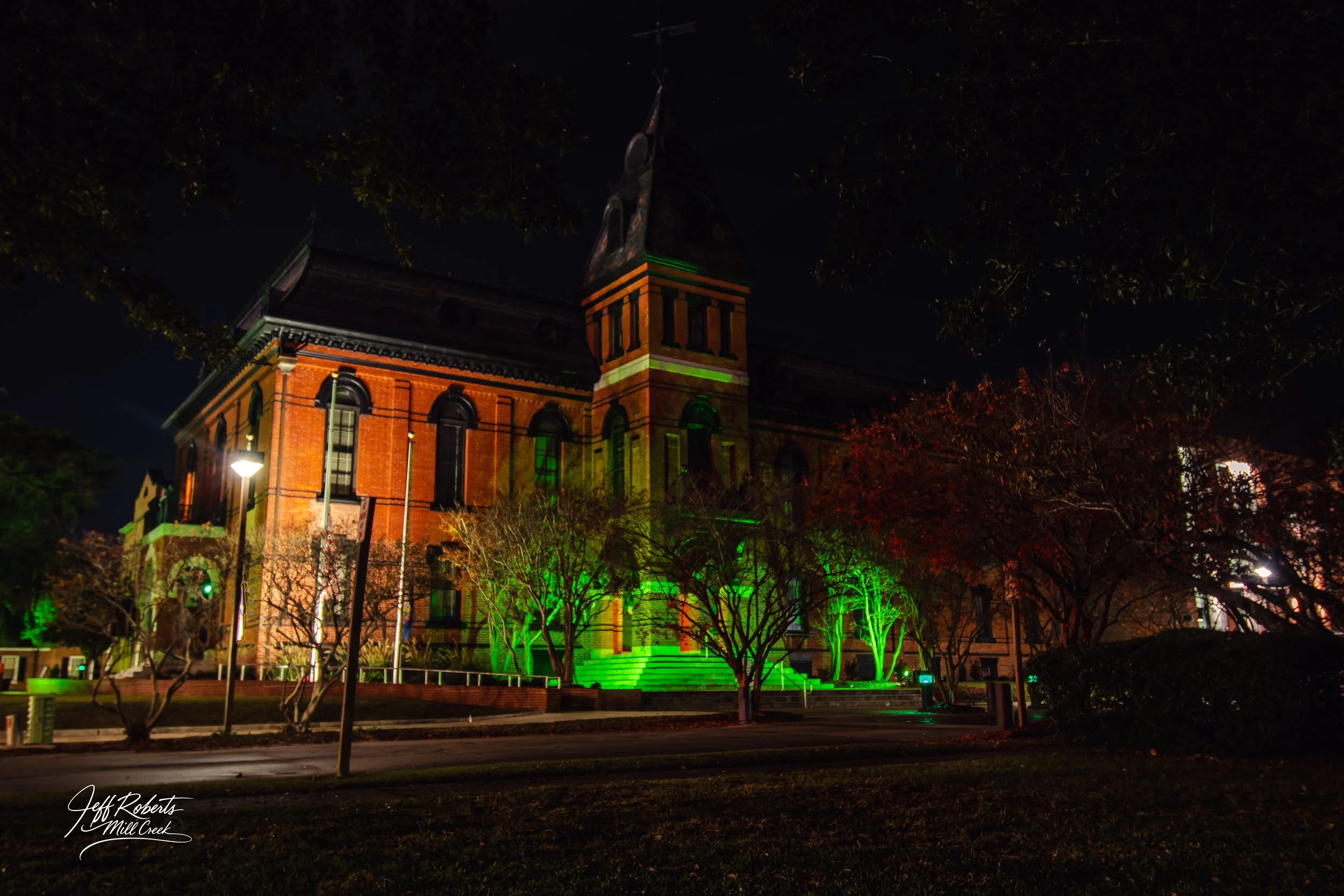 Night view of a historic brick building illuminated with green lights, surrounded by trees and street lamps.