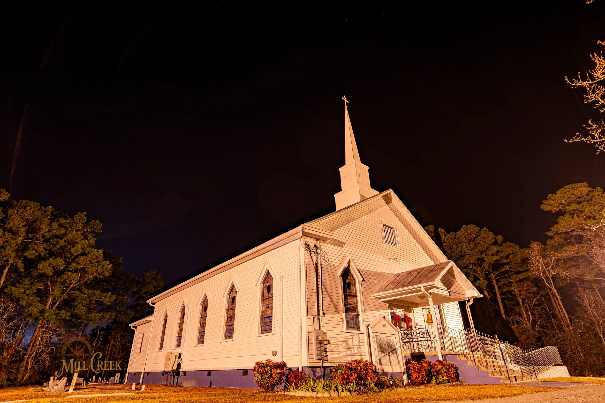 Night view of a white wooden church with tall steeple and cross, illuminated by exterior lighting, surrounded by trees and a landscaped garden