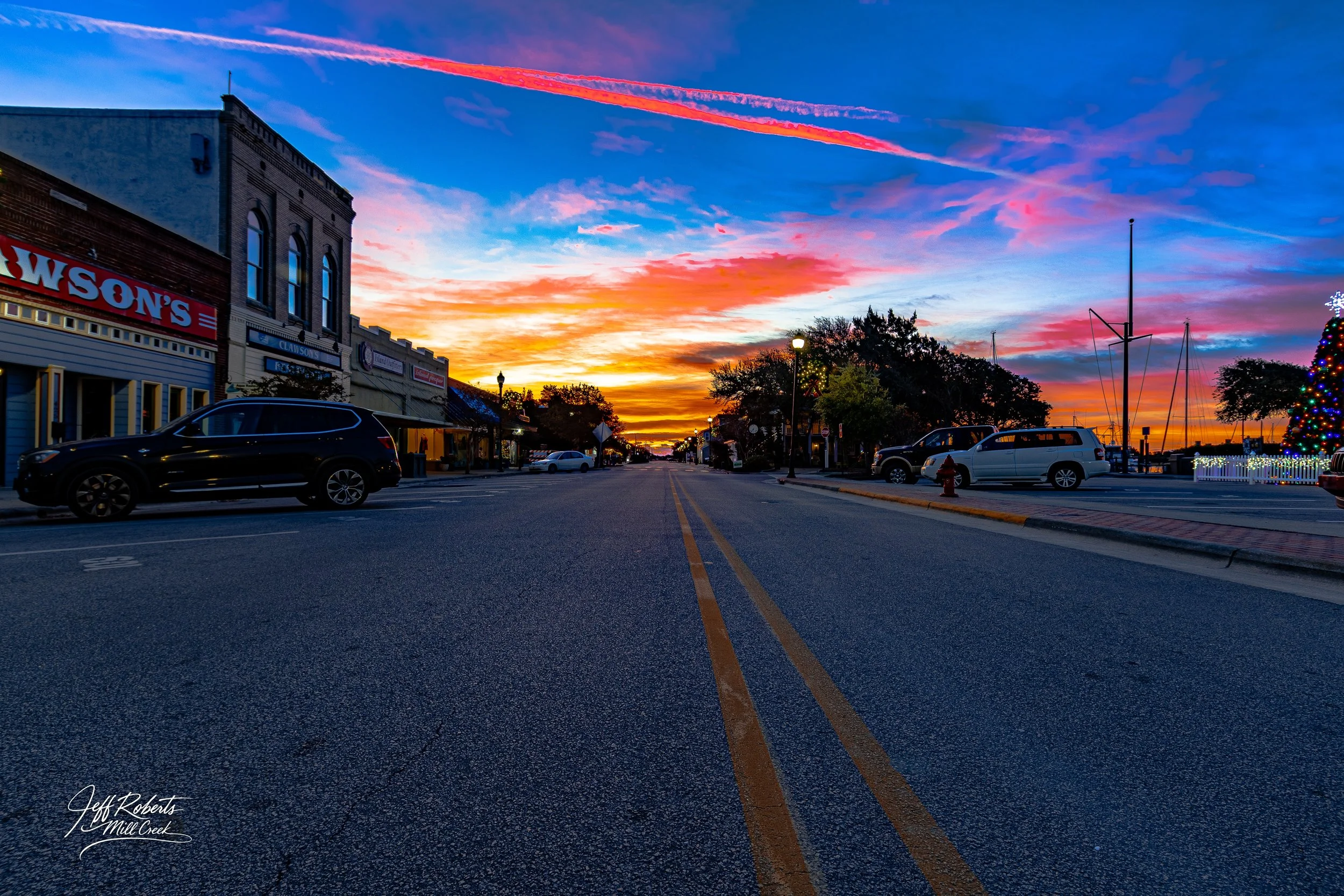 Street view of a small town during sunset with colorful sky, parked cars, shops, Trees, and a decorated Christmas tree on the right.