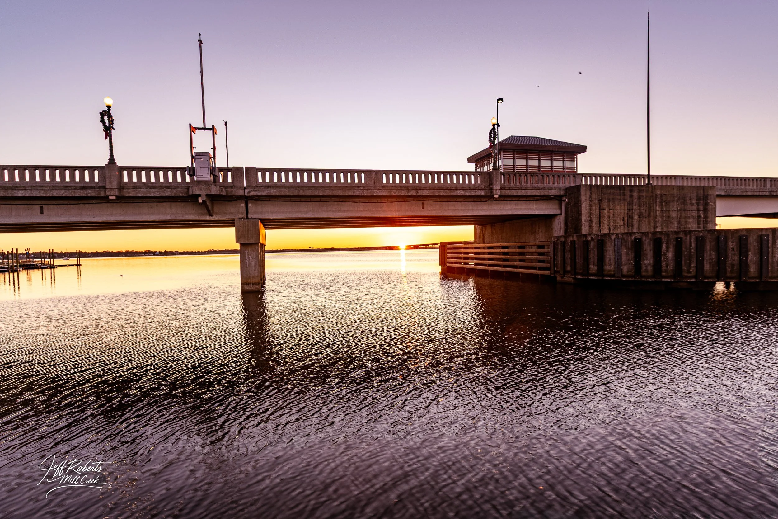 A bridge over a body of water at sunset with the sun visible on the horizon and the sky transitioning from blue to orange. The water reflects the sky and bridge.