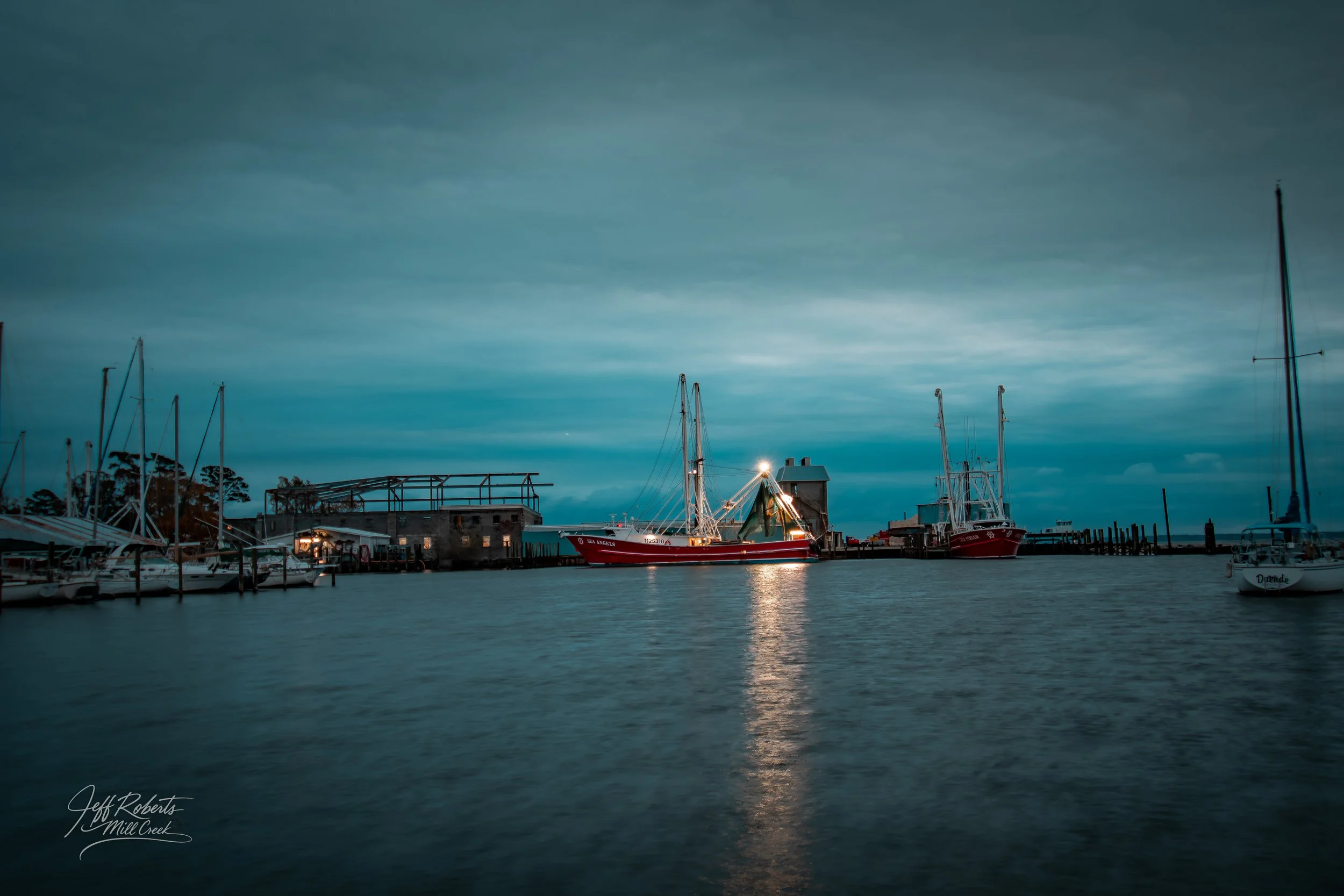 View of a marina with several boats docked, a partly cloudy sky, and reflections on the water during dusk.