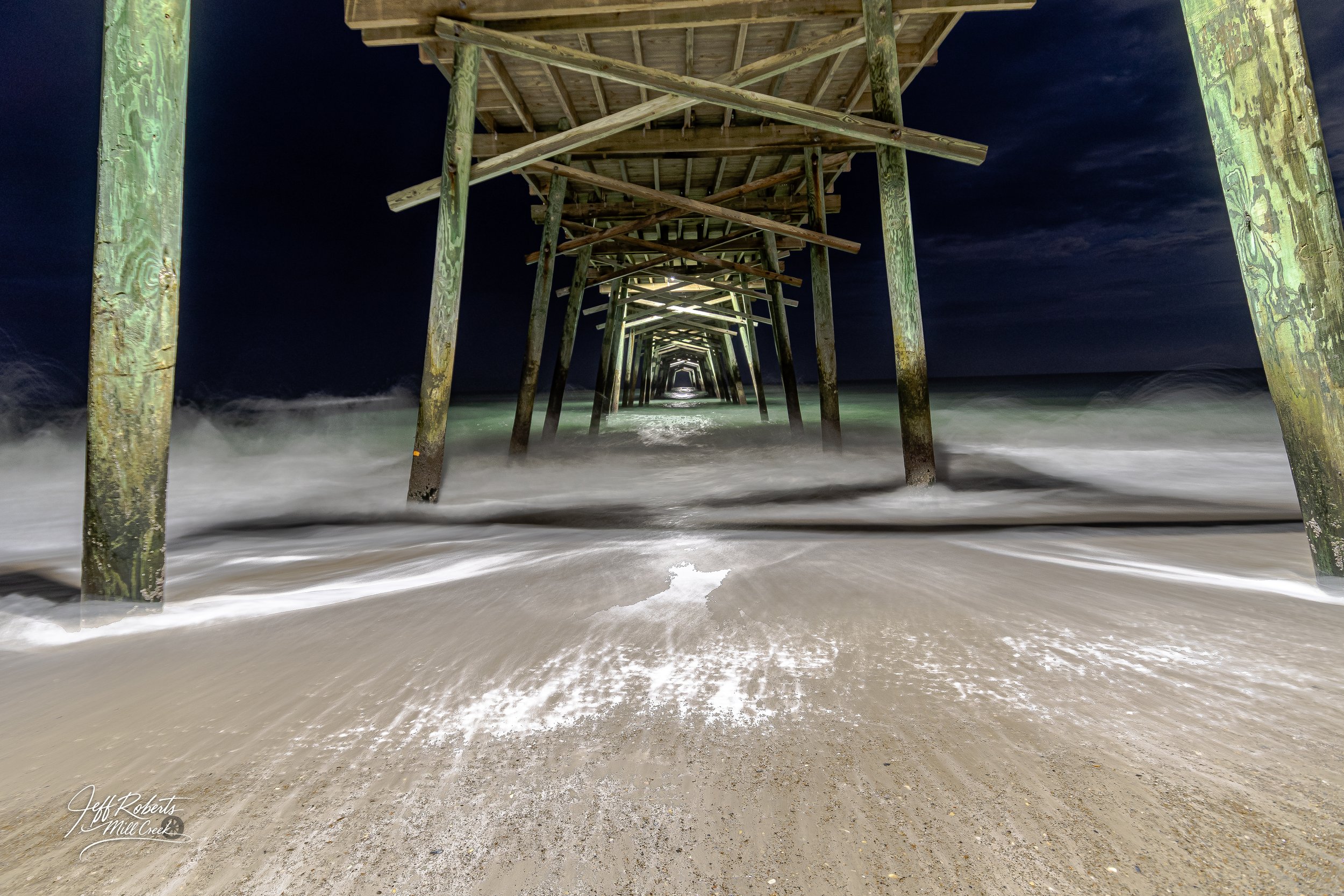 Night view of a wooden pier extending into the ocean with waves washing onto the sandy shore, under a dark sky.