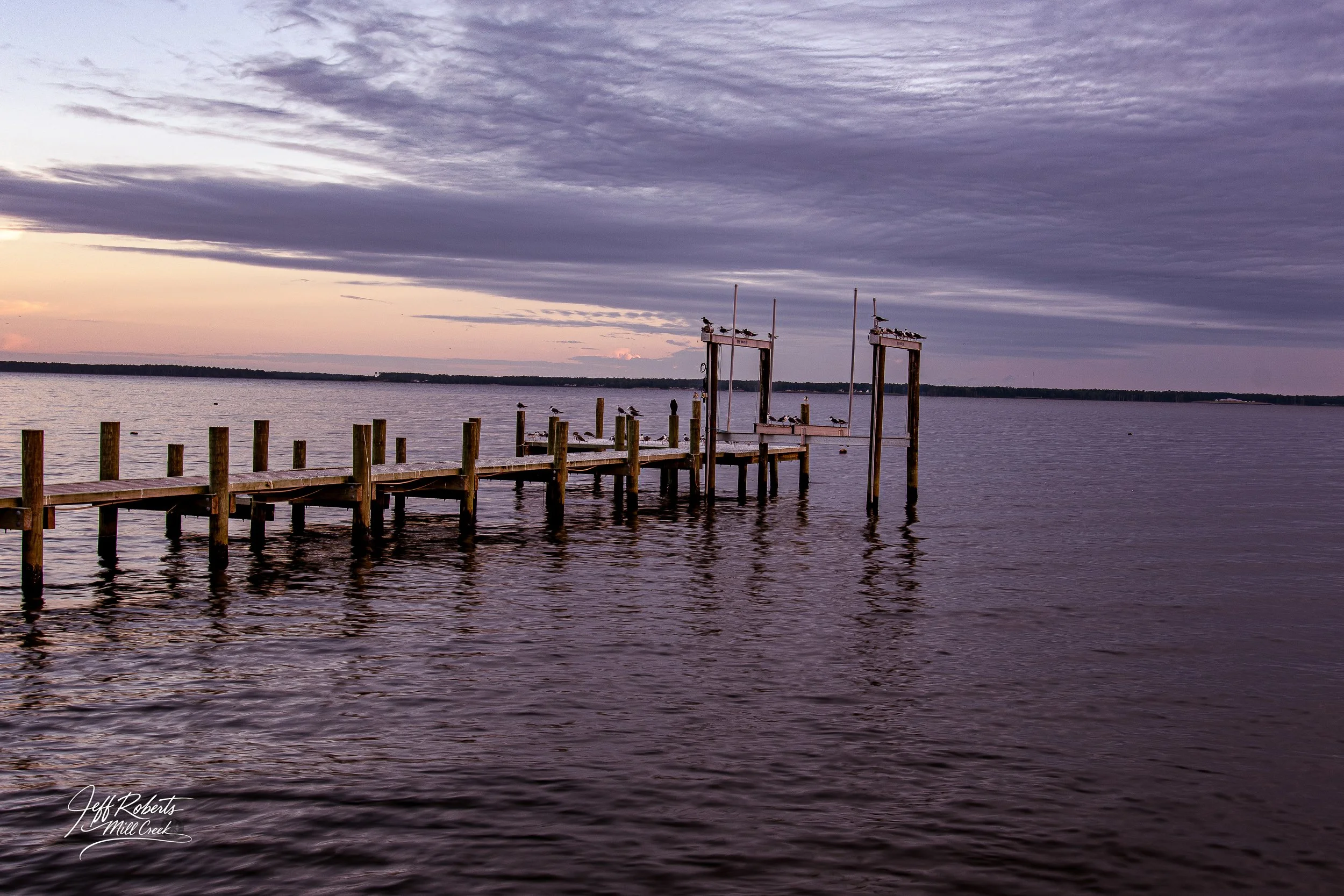 A wooden dock extending into a calm body of water with seagulls perched on the posts and two structures at the end. The sky is partly cloudy with a purple hue during sunset or sunrise.