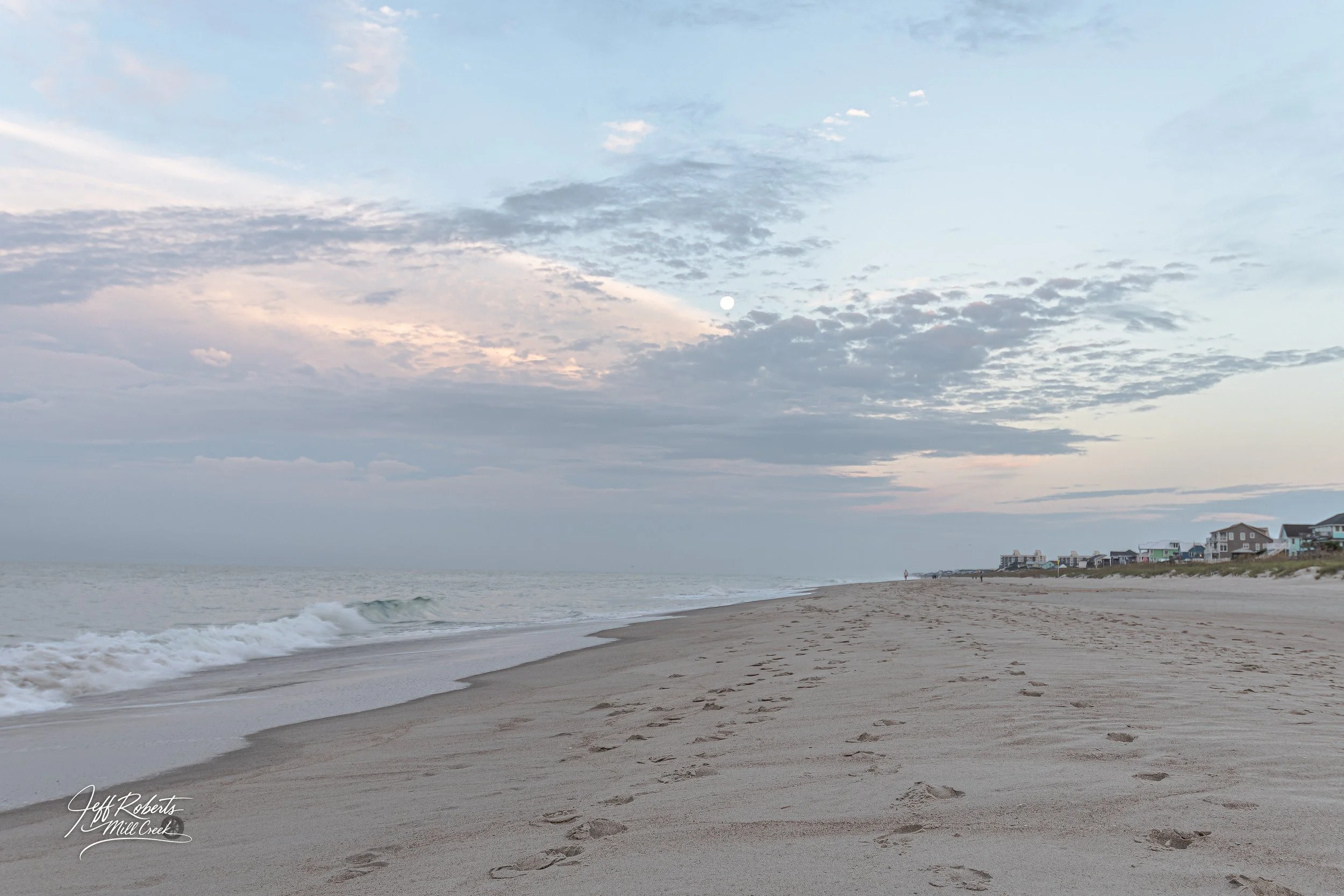 Calm beach with footprints in the sand, gentle waves, and houses in the distance under a partly cloudy sky.