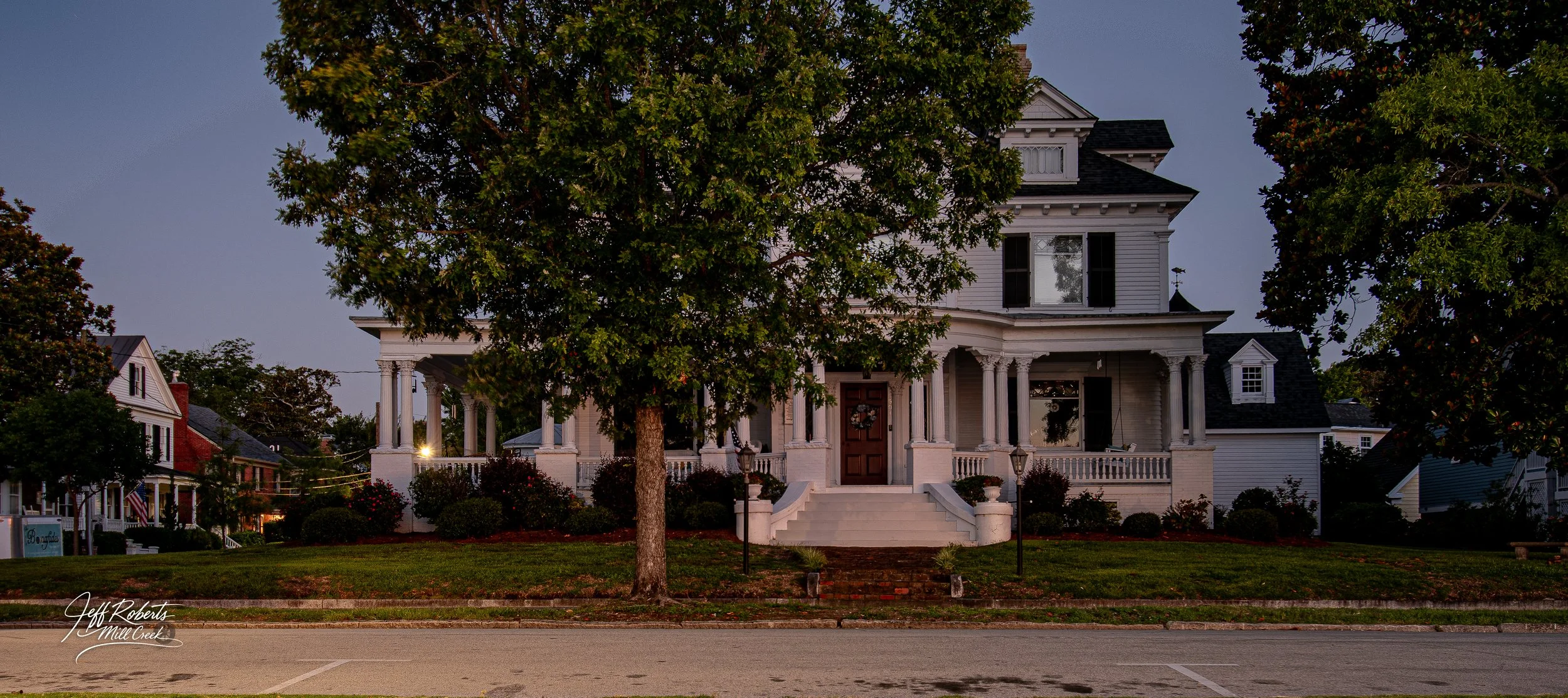 A large, white, Victorian-style house with a front porch, stairs, and multiple stories, partially obscured by two large trees in the front yard, during dusk or early evening.