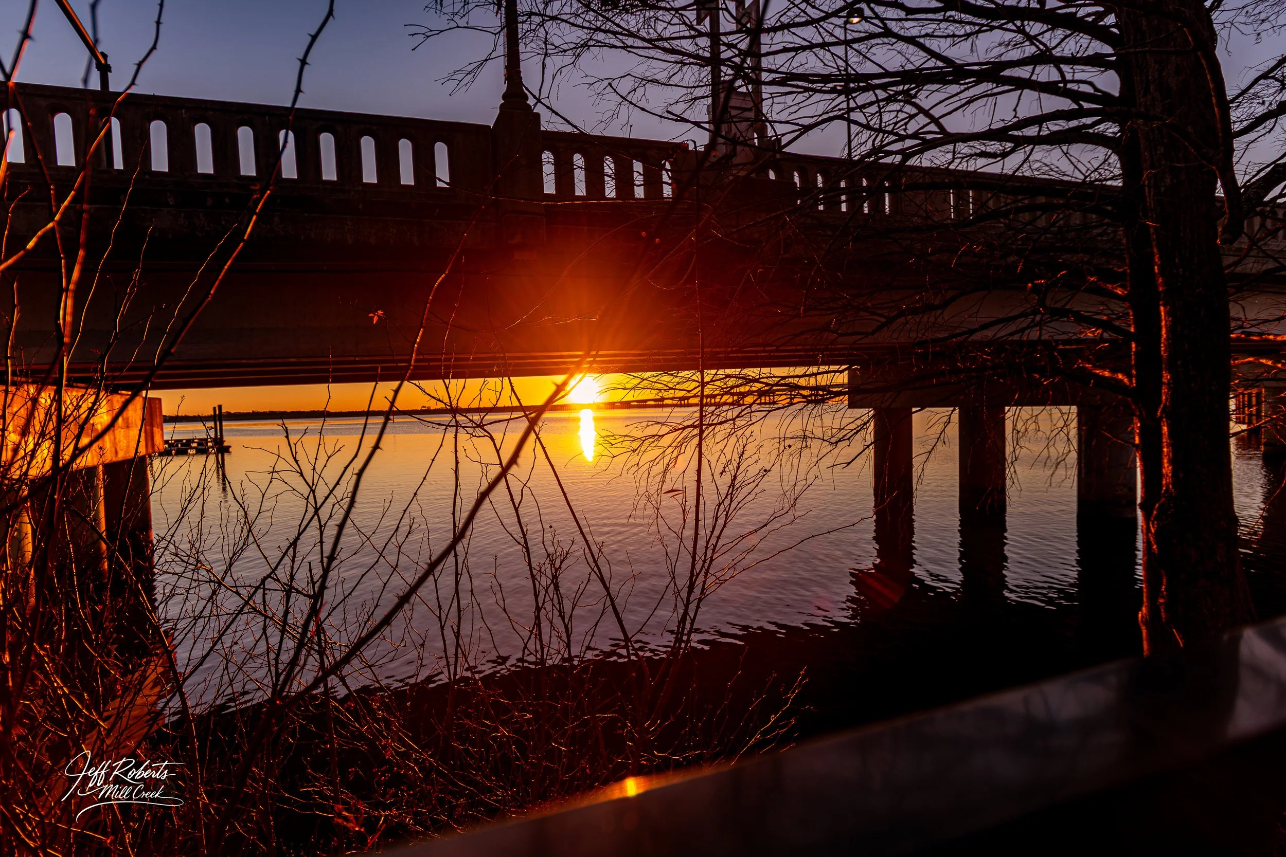 Sunset over a body of water viewed from underneath a bridge, with leafless trees and their branches in the foreground.