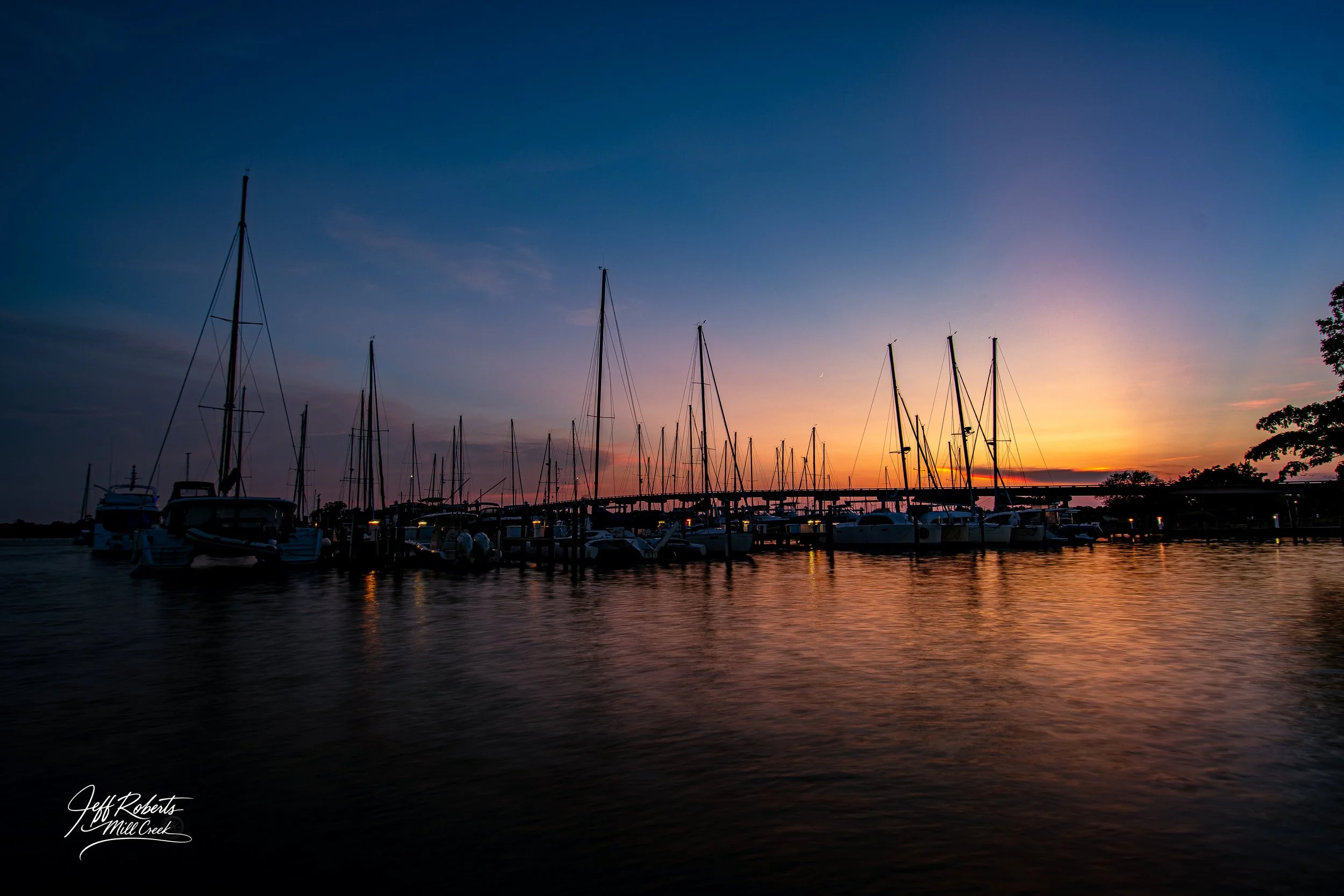 Silhouettes of sailboats docked at a marina during sunset, with the sky transitioning from blue to orange and reflecting on the water.