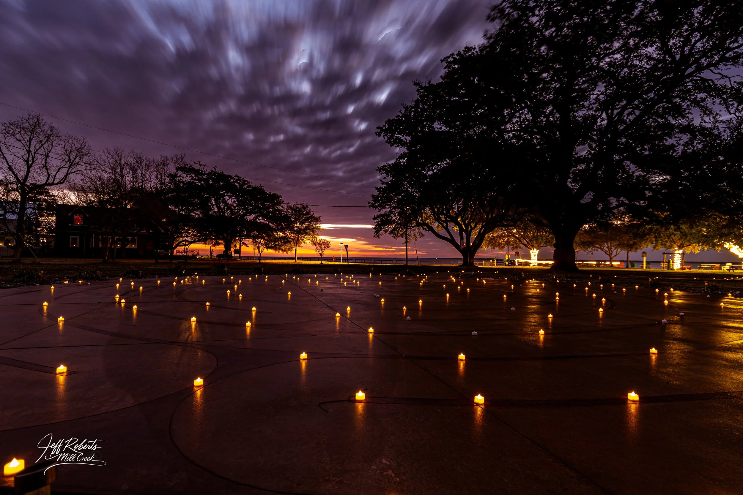 A park with numerous small candle lights on the ground, large silhouetted trees, a cloudy purple sky at sunset, and some holiday lights in the background.