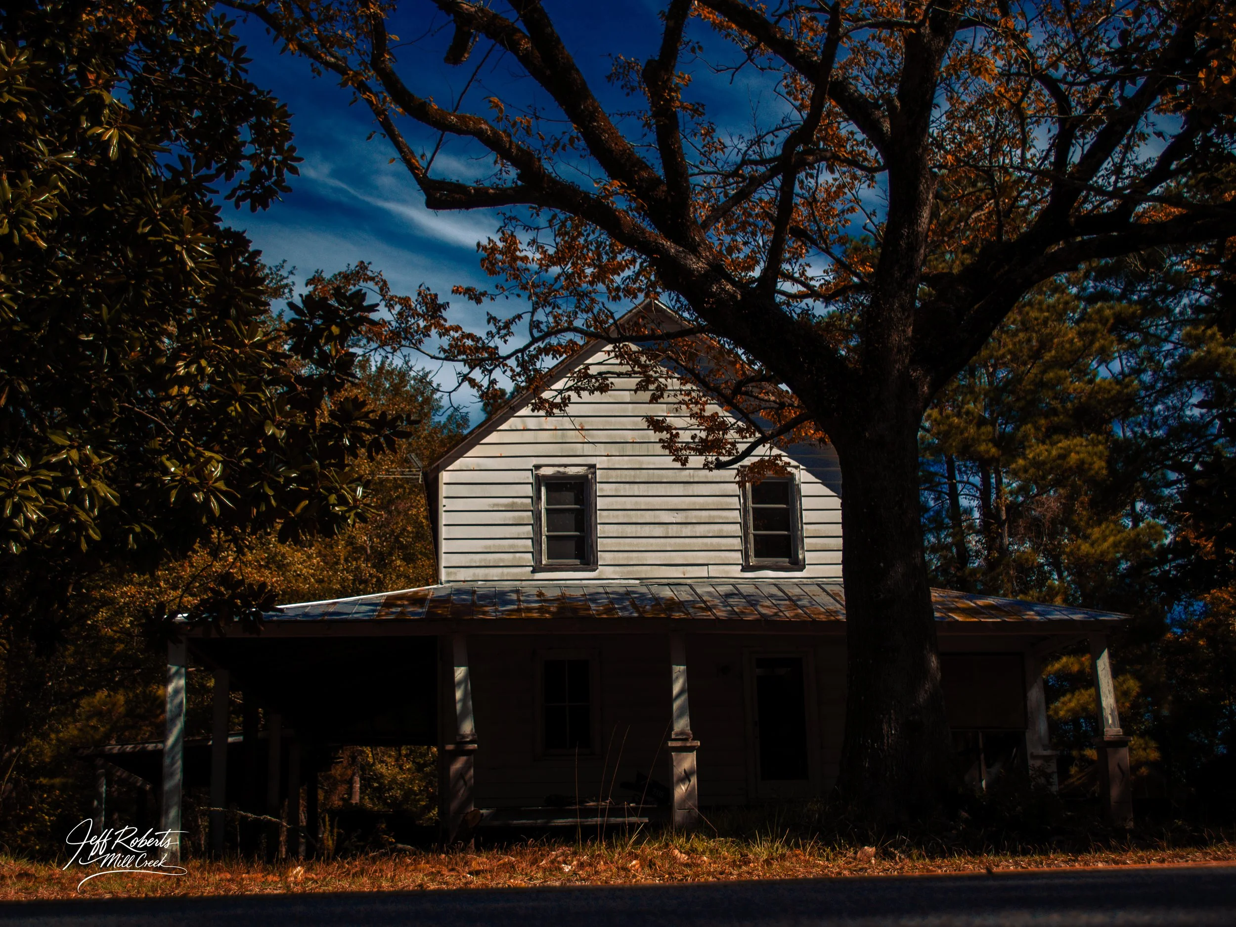 Old white wooden house with two windows on the upper floor, surrounded by trees with autumn foliage, under a clear blue sky.